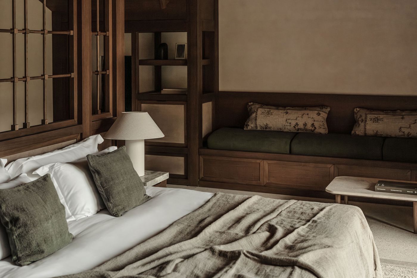 This bedroom exudes a sophisticated rustic minimalism, featuring rich timber paneling and custom built-in furniture. A plush bed with linen throws and olive-green cushions complements a banquette seating area with patterned pillows. Subtle lighting from a modern table lamp illuminates the warm, inviting atmosphere, reflecting a serene and authentic design. This bedroom exudes a sophisticated rustic minimalism, featuring rich timber paneling and custom built-in furniture. A plush bed with linen throws and olive-green cushions complements a banquette seating area with patterned pillows. Subtle lighting from a modern table lamp illuminates the warm, inviting atmosphere, reflecting a serene and authentic design.