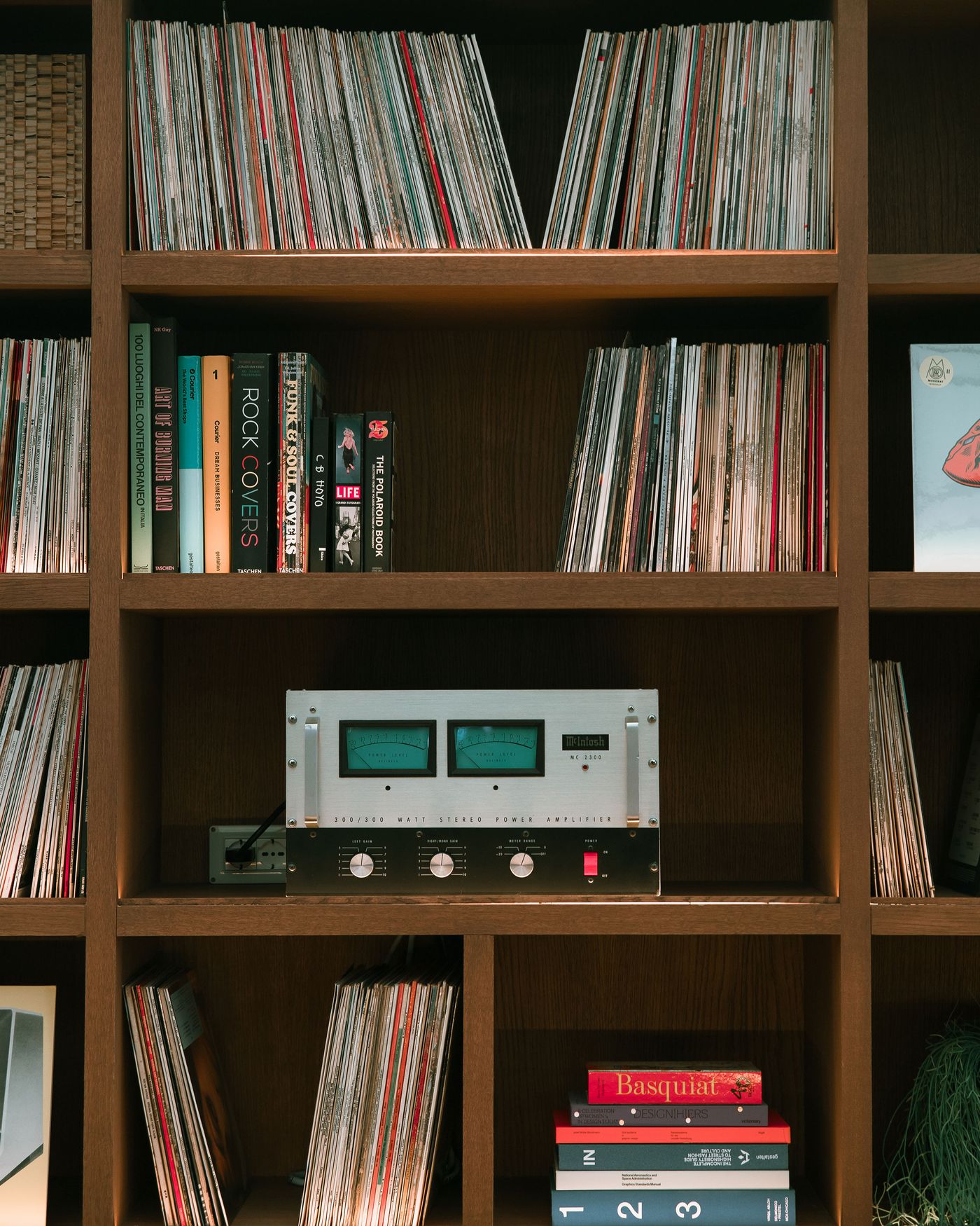 A wooden shelf filled with vinyl records and colorful books, featuring a vintage McIntosh stereo amplifier at the center.