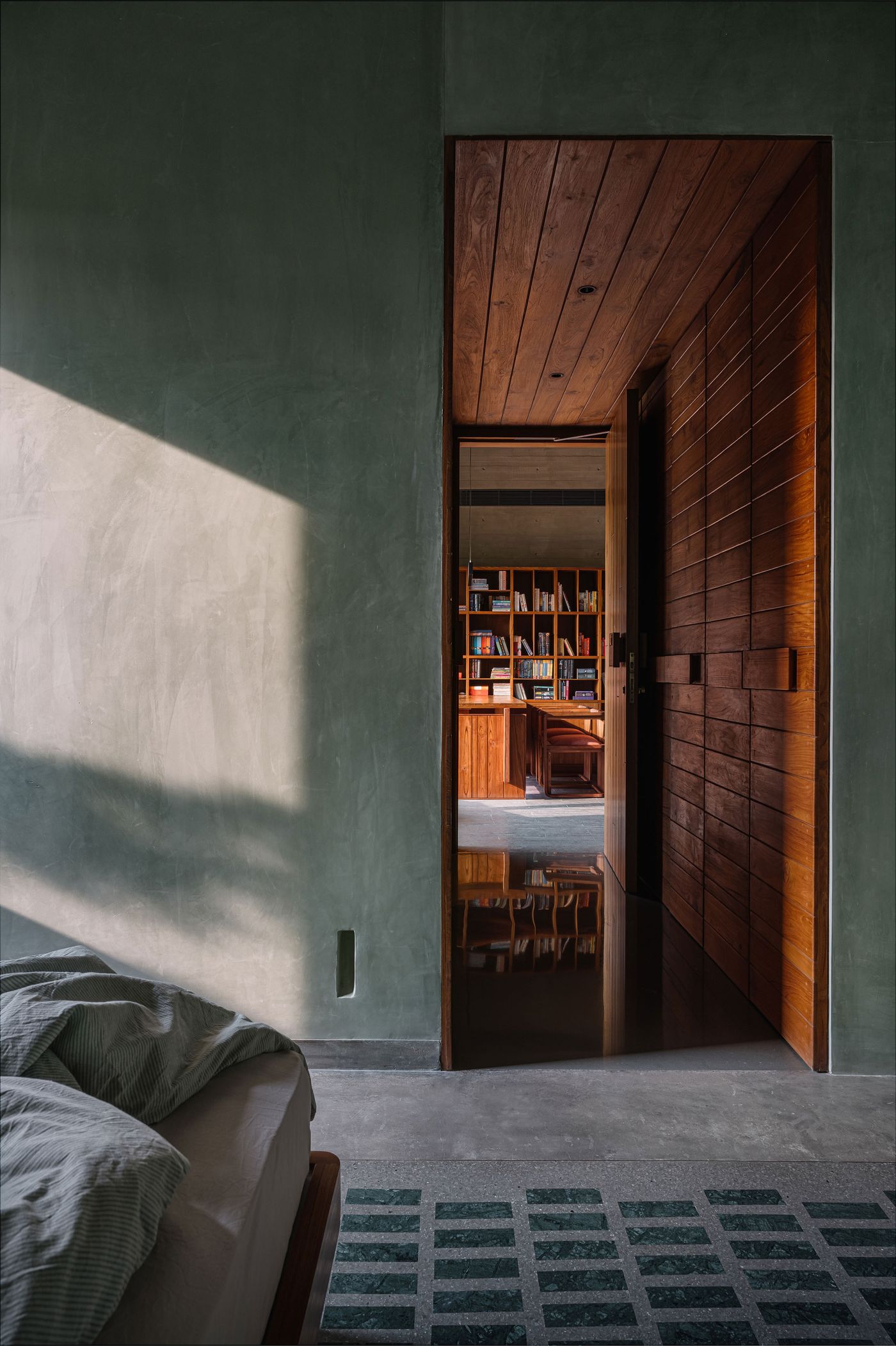 A glimpse into a study or library through an open wooden door, revealing a room with warm wooden walls and built-in bookshelves filled with books. Sunlight streams into the foreground, illuminating a textured wall and a dark tiled floor. The doorway acts as a frame, emphasizing the inviting atmosphere of the room beyond.