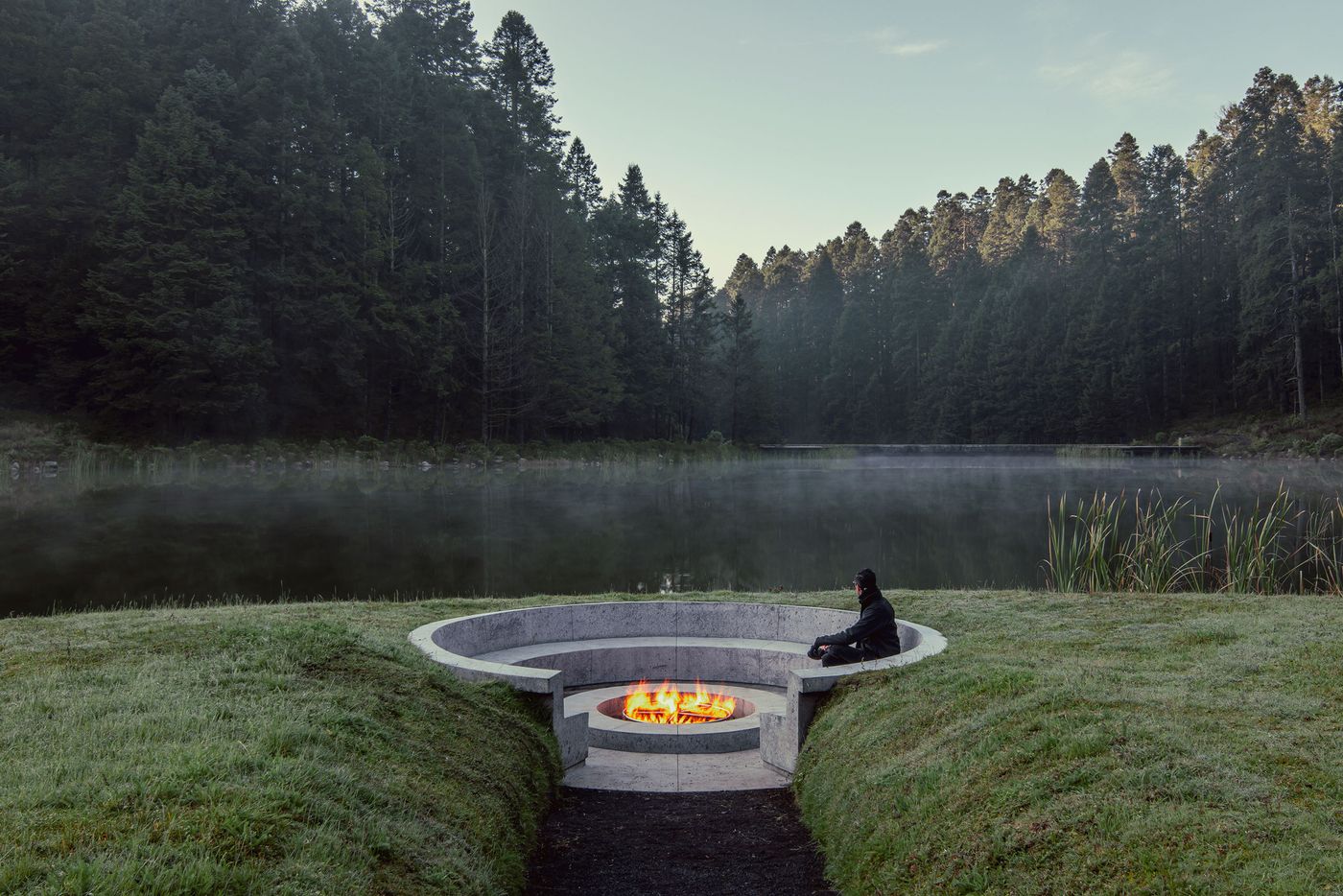 A serene lake setting with mist, featuring a person sitting by a circular concrete firepit amidst lush green grass and tall trees.