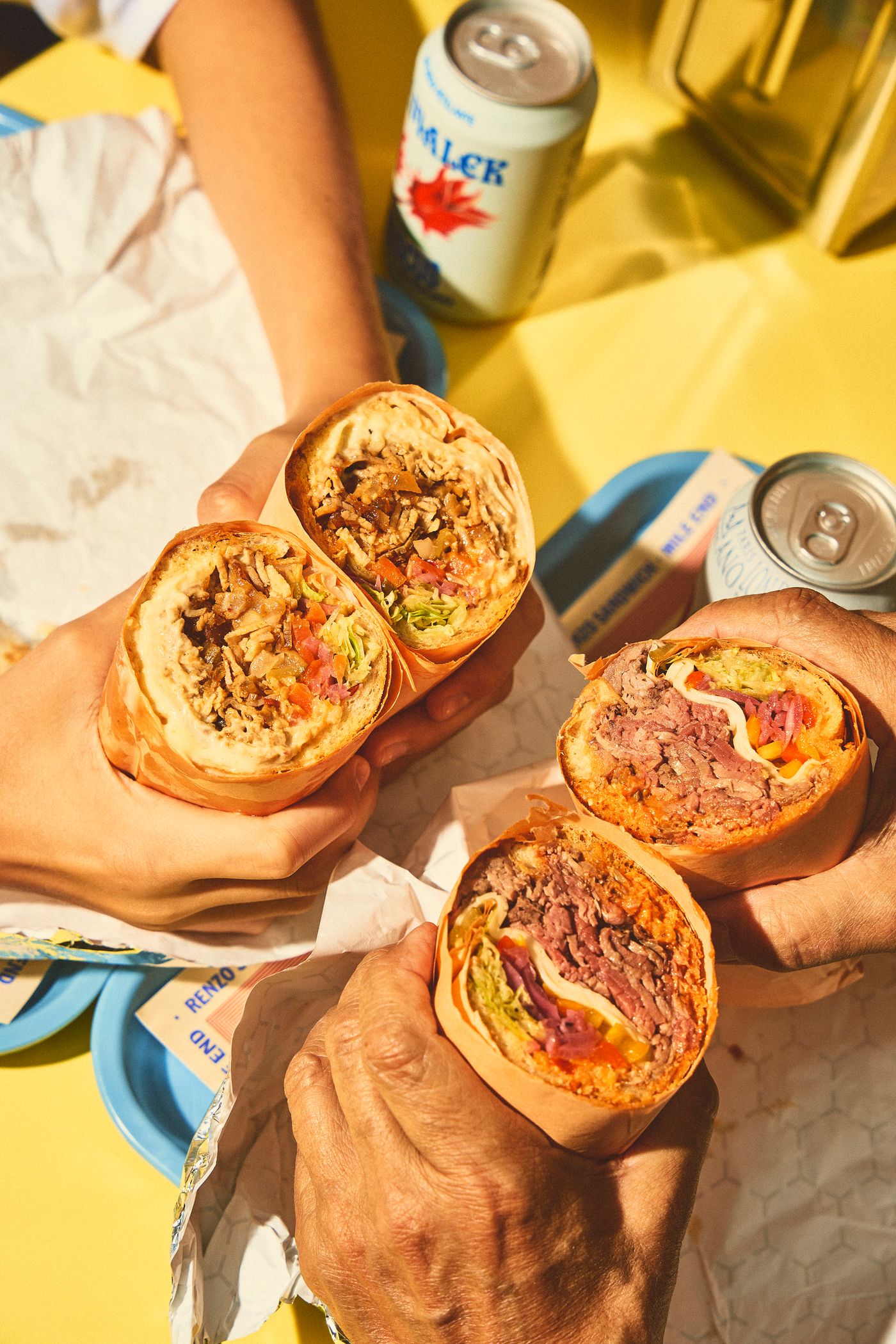 Several hands hold generously filled sandwiches wrapped in paper. Two cans, one with a maple leaf design and another branded "RENZO," sit on a bright yellow surface. The close-up emphasizes the food's textures and ingredients, highlighting the hearty, satisfying nature of the sandwiches.