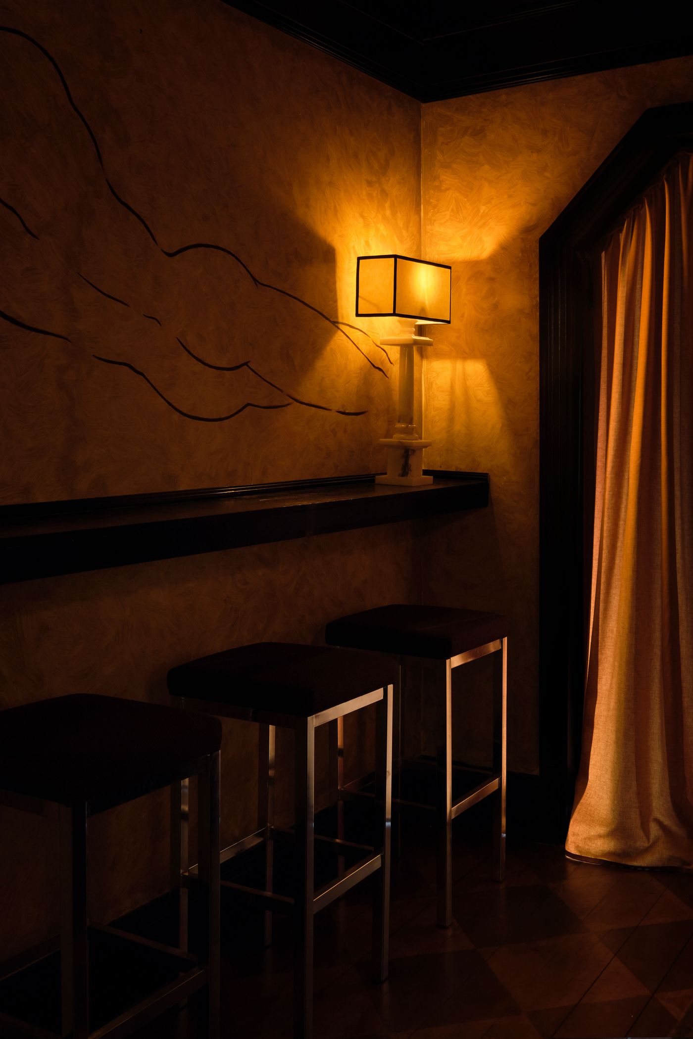 A captivating corner in The Roses of Elagabalus, featuring a minimalist table lamp with a dark rectangular shade casting a warm glow onto a textured, ochre-hued wall adorned with abstract linear art. A slender dark shelf hovers above two sleek, dark barstools, creating an intimate, art-deco inspired nook with a moody atmosphere.