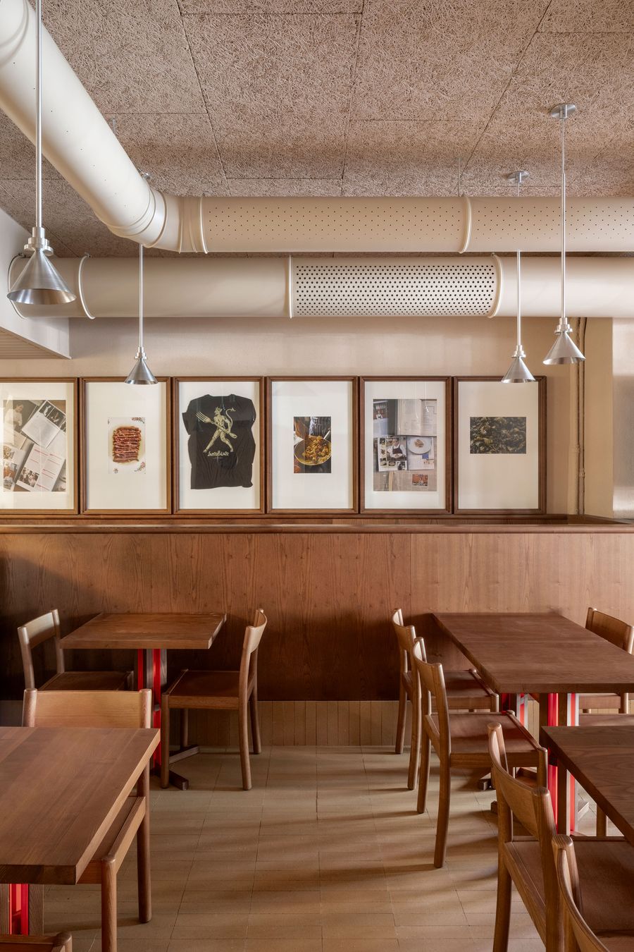 An intimate dining alcove at Santo Palato by Naessi Studio, showcasing the blend of rustic and industrial elements. Framed artworks are precisely aligned above a continuous wooden banquette, complemented by individual wooden tables with red bases. Exposed ceiling ducts and minimalist pendant lights underscore the space's authentic, unpretentious charm. An intimate dining alcove at Santo Palato by Naessi Studio, showcasing the blend of rustic and industrial elements. Framed artworks are precisely aligned above a continuous wooden banquette, complemented by individual wooden tables with red bases. Exposed ceiling ducts and minimalist pendant lights underscore the space's authentic, unpretentious charm.