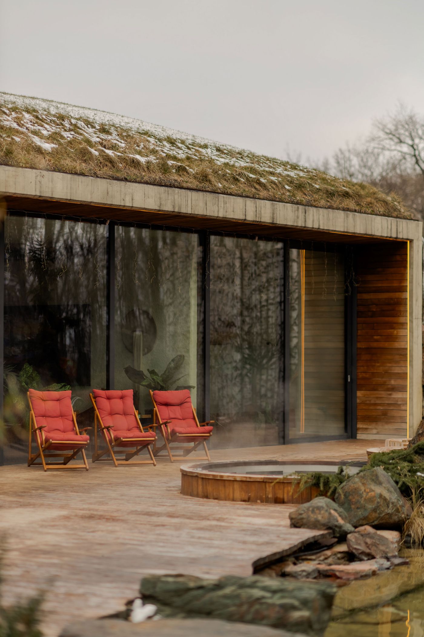 A modern deck features three red lounge chairs, a wooden hot tub, and a green roof, surrounded by a serene natural landscape. A modern deck features three red lounge chairs, a wooden hot tub, and a green roof, surrounded by a serene natural landscape.