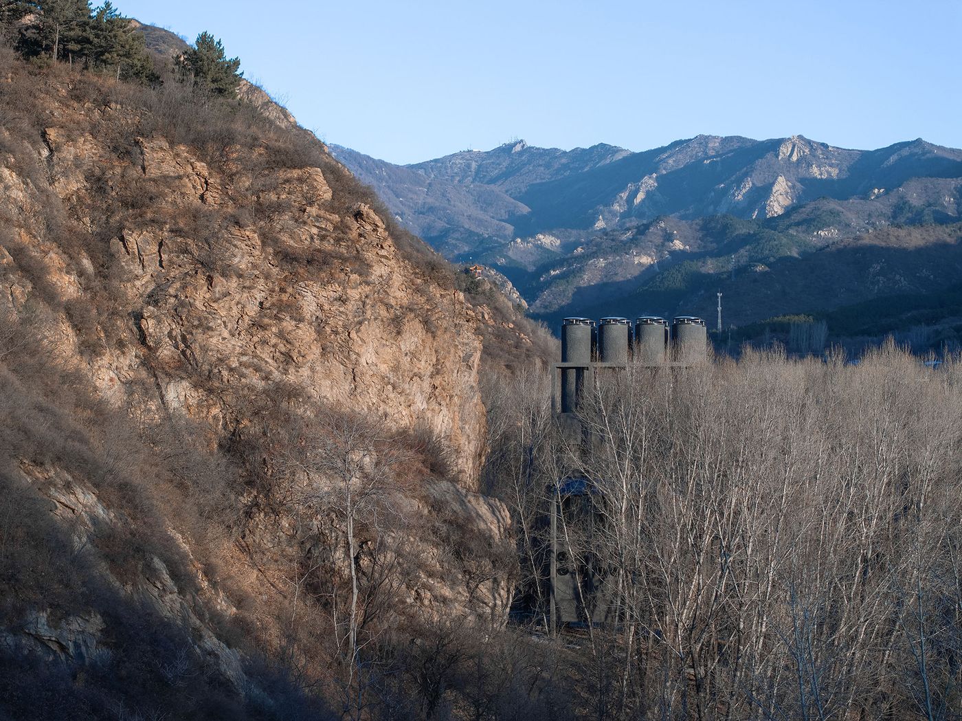 A rocky hillside with sparse trees in the foreground, featuring large industrial silos against a backdrop of distant mountains. A rocky hillside with sparse trees in the foreground, featuring large industrial silos against a backdrop of distant mountains.