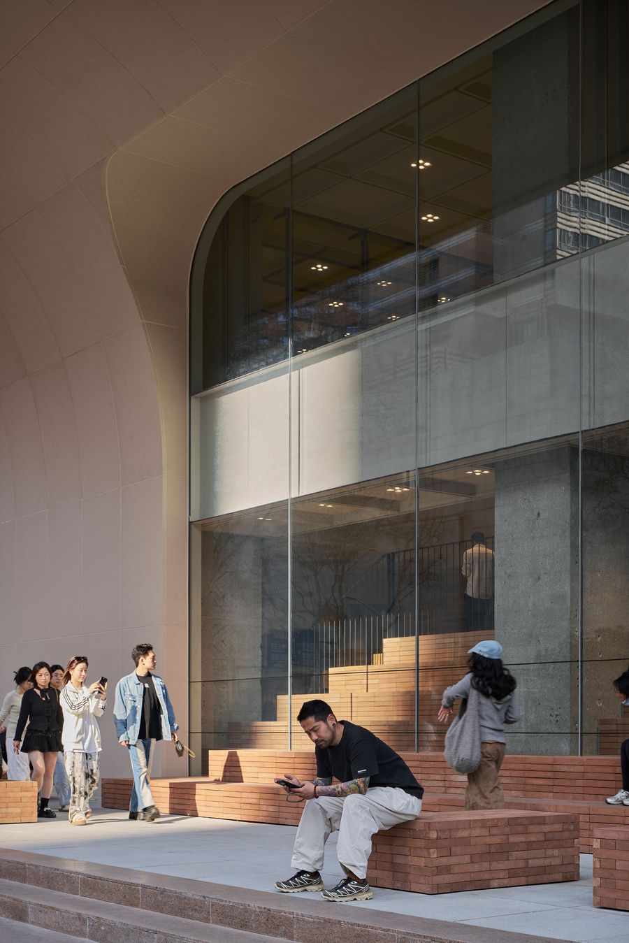 A modern building facade features large glass windows and an elegant archway, with people interacting on brick seating outside. A modern building facade features large glass windows and an elegant archway, with people interacting on brick seating outside.