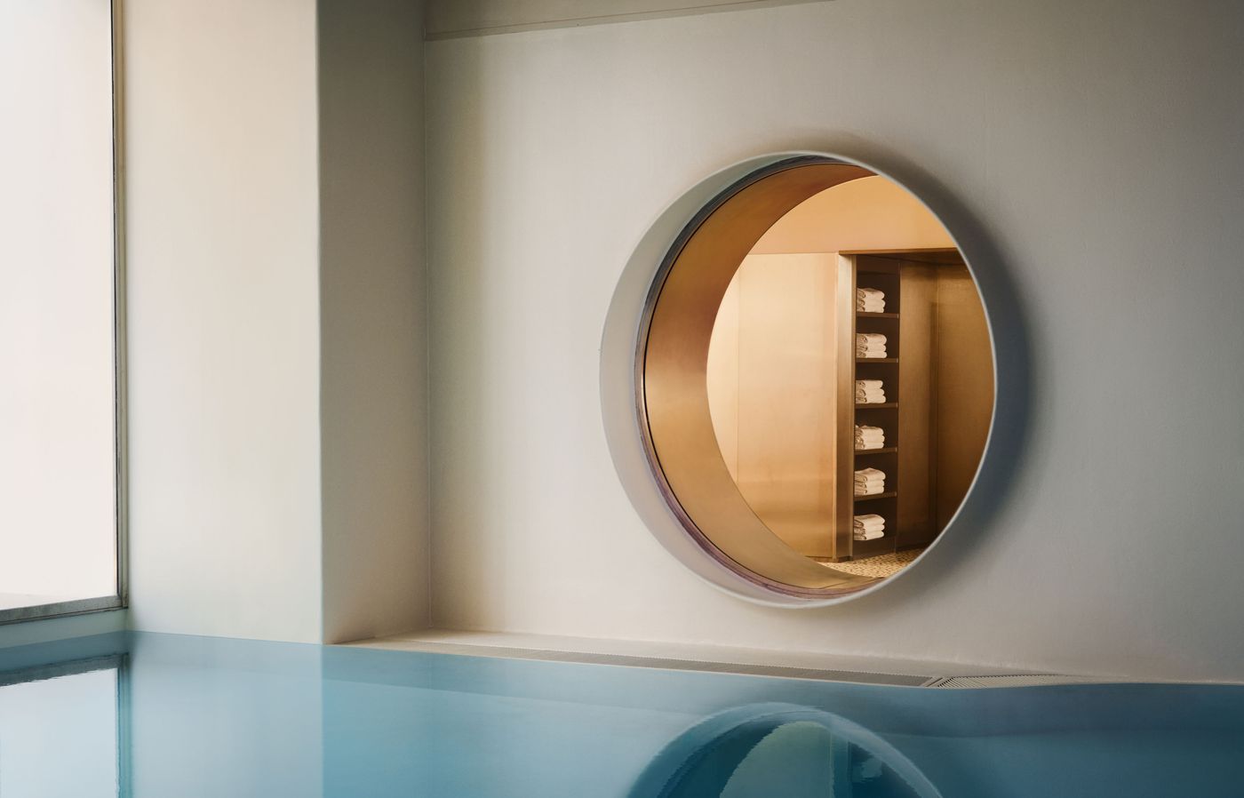 Seen from water level, the Ken Club pool is defined by a large porthole-like opening set into a pale wall, revealing neatly stacked towels beyond. The blue surface mirrors the circular aperture, creating a near-symmetrical composition that emphasises the project’s restrained yet playful geometry. Seen from water level, the Ken Club pool is defined by a large porthole-like opening set into a pale wall, revealing neatly stacked towels beyond. The blue surface mirrors the circular aperture, creating a near-symmetrical composition that emphasises the project’s restrained yet playful geometry.
