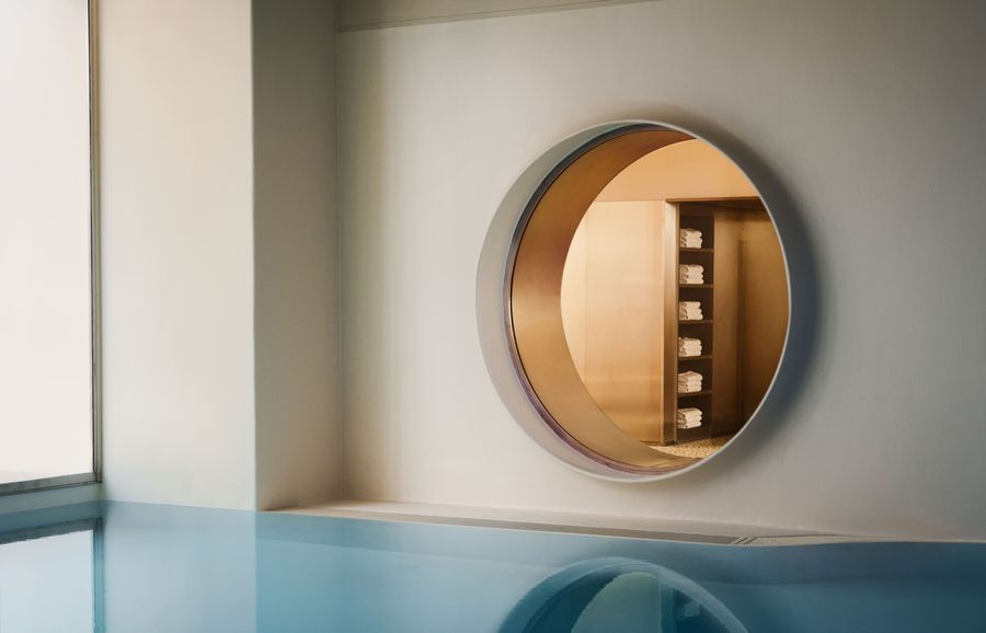 Seen from water level, the Ken Club pool is defined by a large porthole-like opening set into a pale wall, revealing neatly stacked towels beyond. The blue surface mirrors the circular aperture, creating a near-symmetrical composition that emphasises the project’s restrained yet playful geometry.