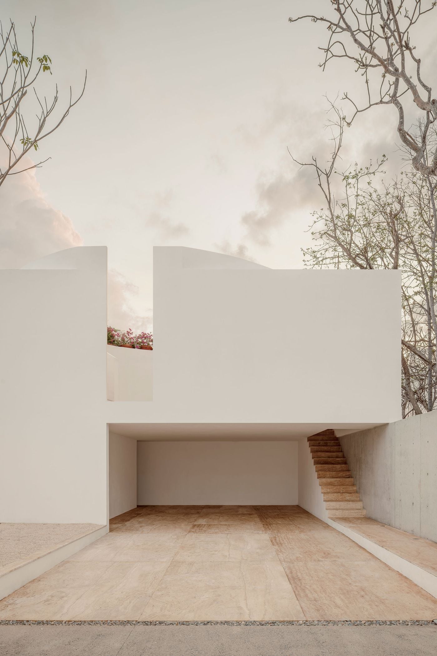 Beyond the street gates, a deep covered forecourt opens beneath the white upper volume, its warm veined paving flowing into a floating stair that climbs alongside a concrete wall. Above, a slot between the masses reveals potted pink blossoms, while gently curving parapets hint at the courtyard within. Beyond the street gates, a deep covered forecourt opens beneath the white upper volume, its warm veined paving flowing into a floating stair that climbs alongside a concrete wall. Above, a slot between the masses reveals potted pink blossoms, while gently curving parapets hint at the courtyard within.