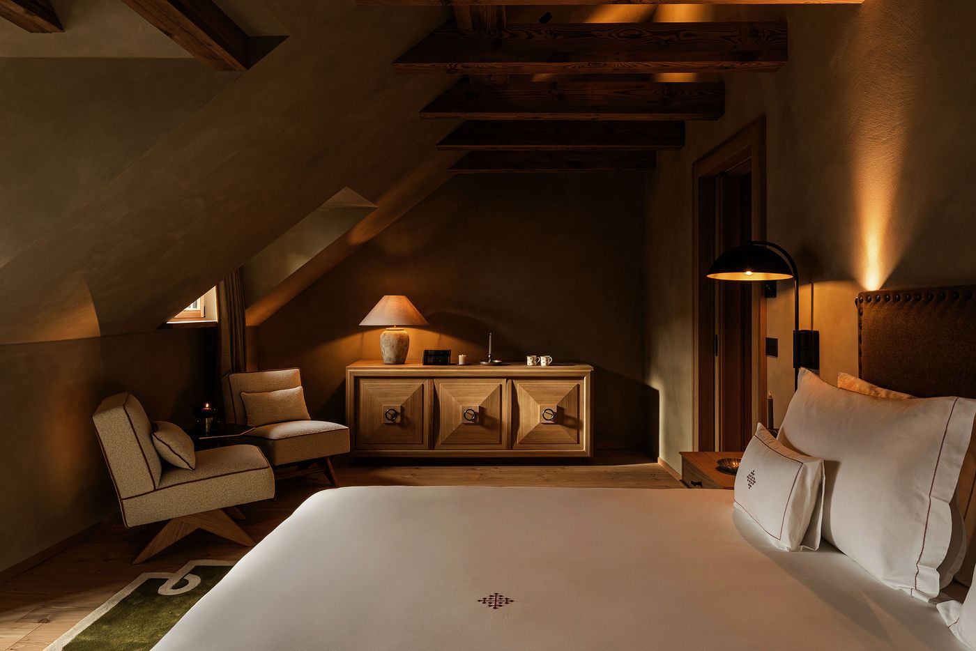 A guest room in the eaves of Castel Badia pairs dark timber beams and sloping plastered walls with a diamond-point oak sideboard and two bouclé slipper chairs on angled wooden legs. A domed reading lamp, a studded upholstered headboard and white linens embroidered with a burgundy cross motif complete the scene. A guest room in the eaves of Castel Badia pairs dark timber beams and sloping plastered walls with a diamond-point oak sideboard and two bouclé slipper chairs on angled wooden legs. A domed reading lamp, a studded upholstered headboard and white linens embroidered with a burgundy cross motif complete the scene.