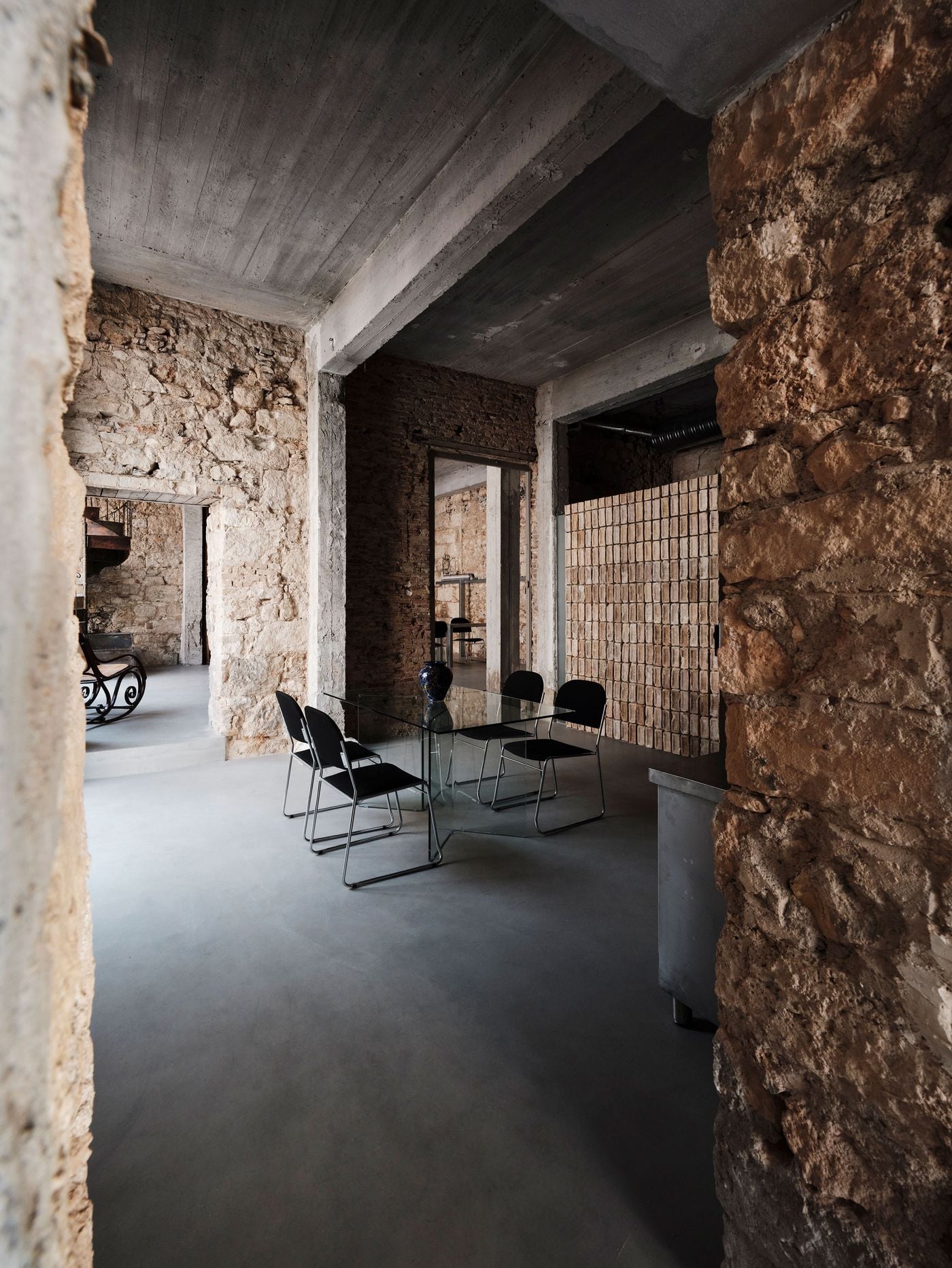 The meeting area unfolds between massive stone walls and exposed concrete beams, with a transparent glass table and lightweight black chairs keeping sightlines open across the interior. A reclaimed-brick partition adds another material layer, while adjoining rooms remain visually connected through the reopened original doorways. The meeting area unfolds between massive stone walls and exposed concrete beams, with a transparent glass table and lightweight black chairs keeping sightlines open across the interior. A reclaimed-brick partition adds another material layer, while adjoining rooms remain visually connected through the reopened original doorways.
