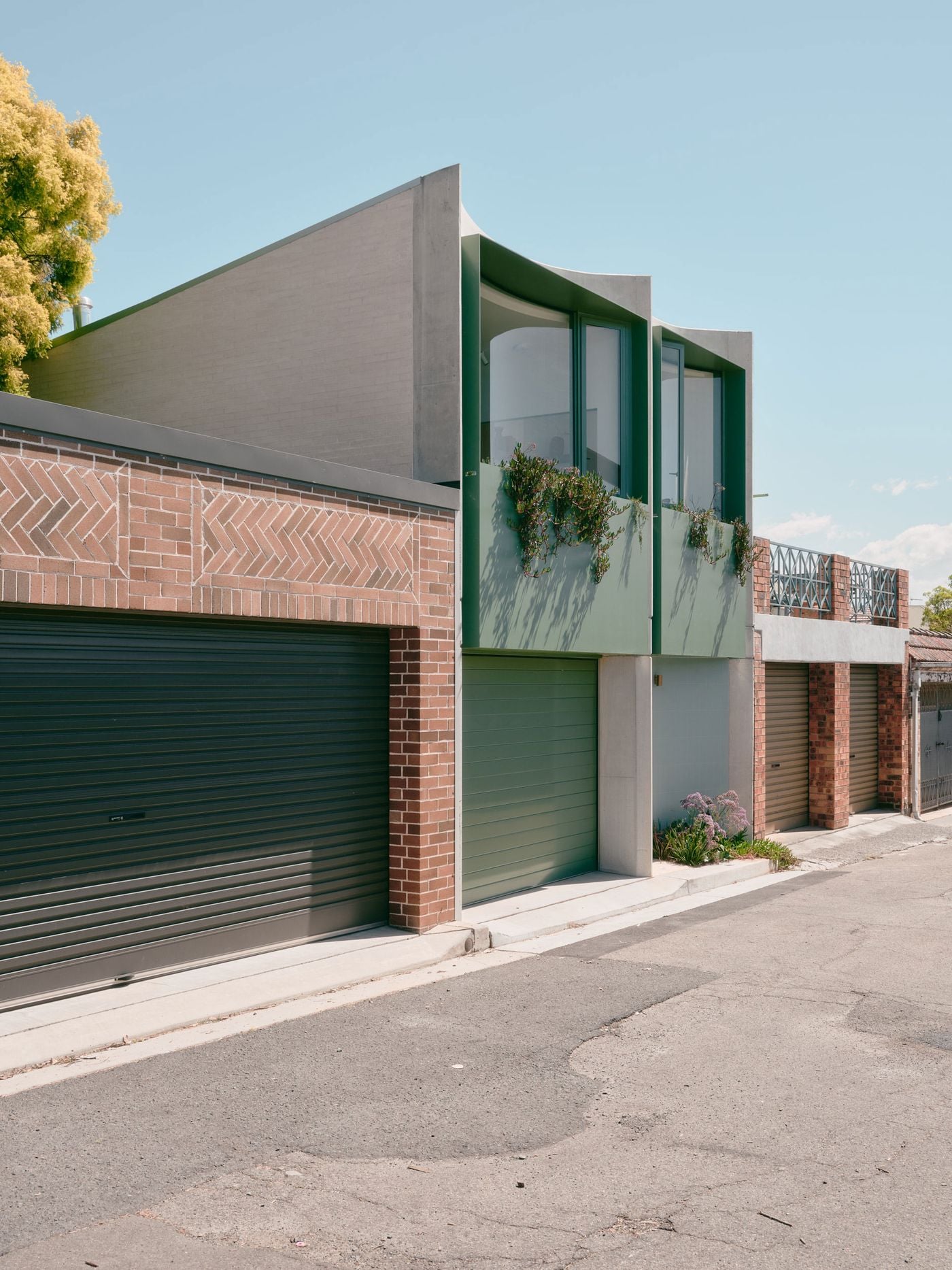 This wider laneway view places the project within its immediate context of garage doors, brick walls and hard paving. Denj Lane breaks the repetitive service-edge condition with a taller, inhabited façade, combining pale brick, sage-green panels, projecting glazing and cascading planting to introduce colour, depth and domestic presence. This wider laneway view places the project within its immediate context of garage doors, brick walls and hard paving. Denj Lane breaks the repetitive service-edge condition with a taller, inhabited façade, combining pale brick, sage-green panels, projecting glazing and cascading planting to introduce colour, depth and domestic presence.