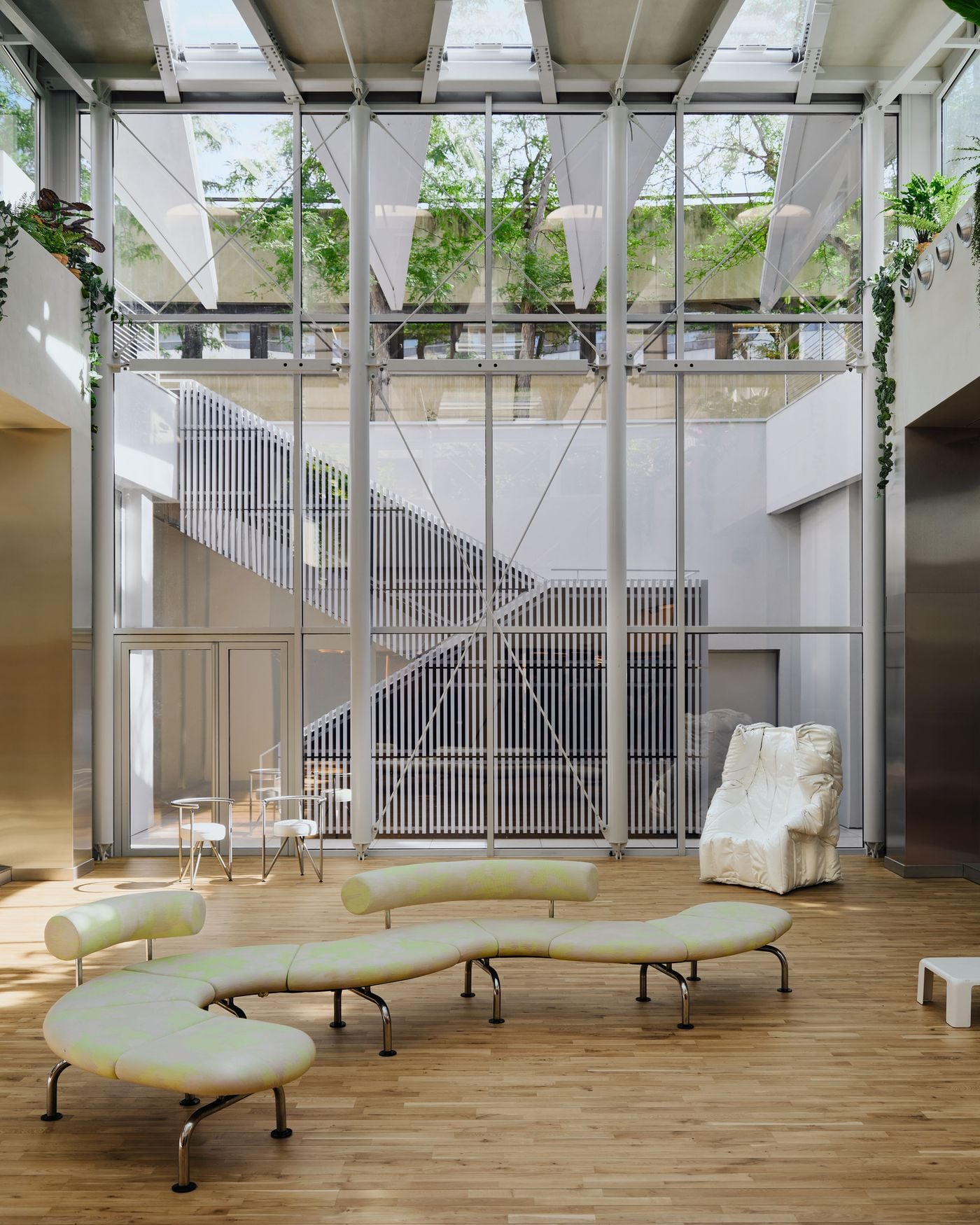The new lounge patio, designed by architect Denis Valode, is enclosed by a soaring glazed structure with exposed white steel framing. A serpentine upholstered bench anchors the timber-floored space, joined by lightweight metal seating and a sculptural white armchair, while daylight and surrounding greenery lend the room an airy, almost conservatory-like atmosphere. The new lounge patio, designed by architect Denis Valode, is enclosed by a soaring glazed structure with exposed white steel framing. A serpentine upholstered bench anchors the timber-floored space, joined by lightweight metal seating and a sculptural white armchair, while daylight and surrounding greenery lend the room an airy, almost conservatory-like atmosphere.