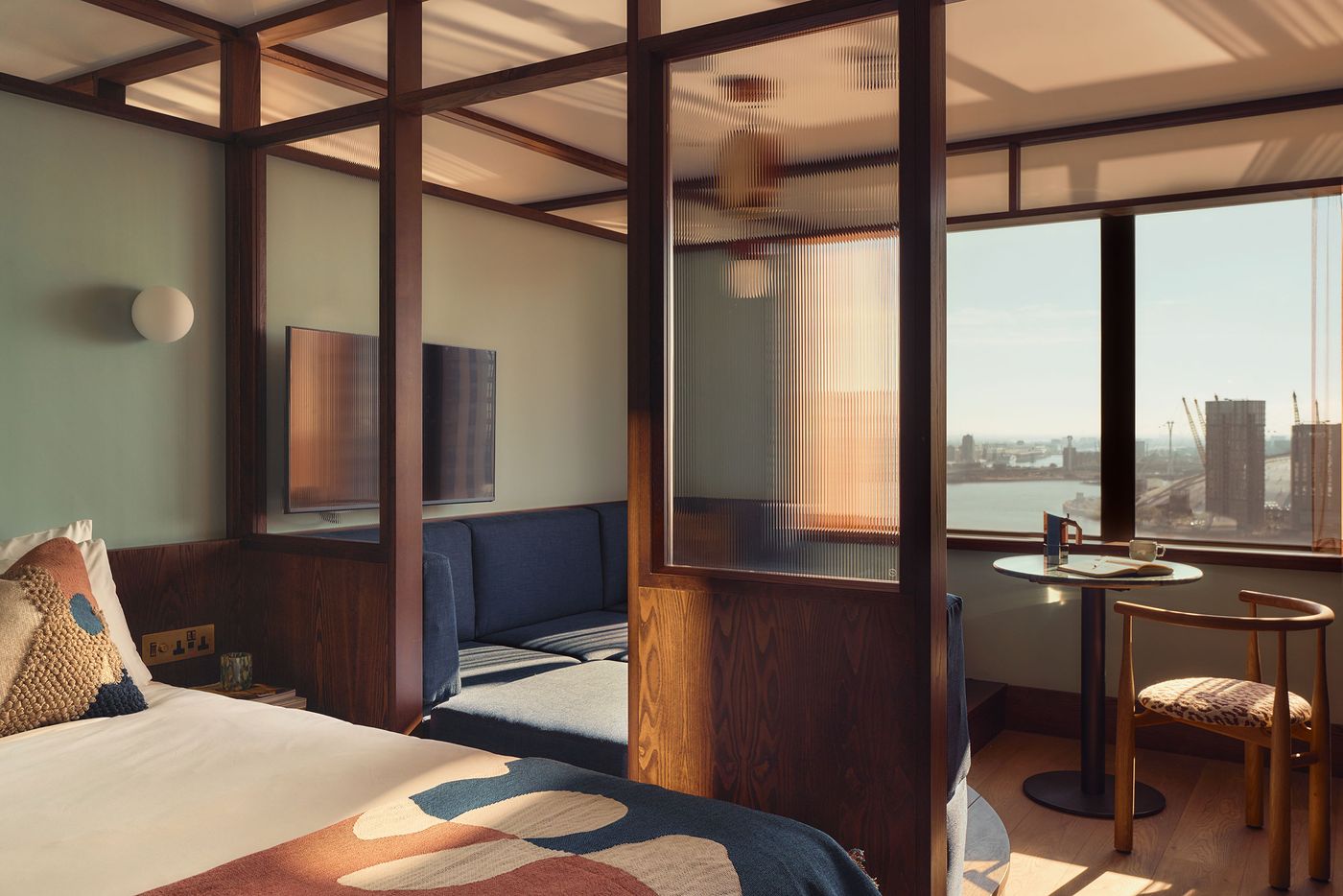 Dark walnut framing gives this Locke London Canary Wharf guest room its distinctly mid-century rhythm, dividing the bed, sofa pit and dining area into interconnected zones. Reeded glass filters sunlight between spaces, while sage walls, blue upholstery and warm timber temper the geometry against expansive views across the Thames. Dark walnut framing gives this Locke London Canary Wharf guest room its distinctly mid-century rhythm, dividing the bed, sofa pit and dining area into interconnected zones. Reeded glass filters sunlight between spaces, while sage walls, blue upholstery and warm timber temper the geometry against expansive views across the Thames.