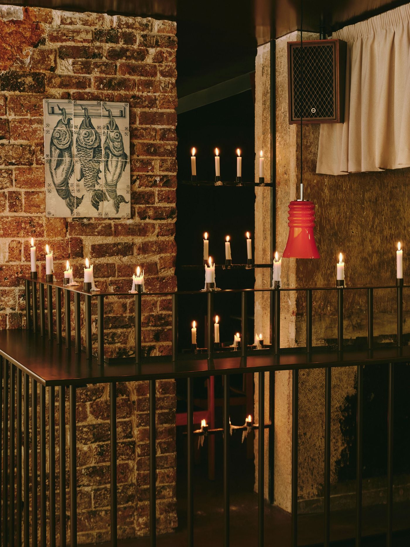 Candles in varying states of meltdown line a dark metal balustrade at SaltoBar in Paris. Wax pools and drips over the slender uprights. Behind, a tiled panel of three hanging fish sits on red-brown brickwork, while a glossy red pendant and a vintage wood-cased speaker add retro colour against rough plaster and gathered linen. Candles in varying states of meltdown line a dark metal balustrade at SaltoBar in Paris. Wax pools and drips over the slender uprights. Behind, a tiled panel of three hanging fish sits on red-brown brickwork, while a glossy red pendant and a vintage wood-cased speaker add retro colour against rough plaster and gathered linen.