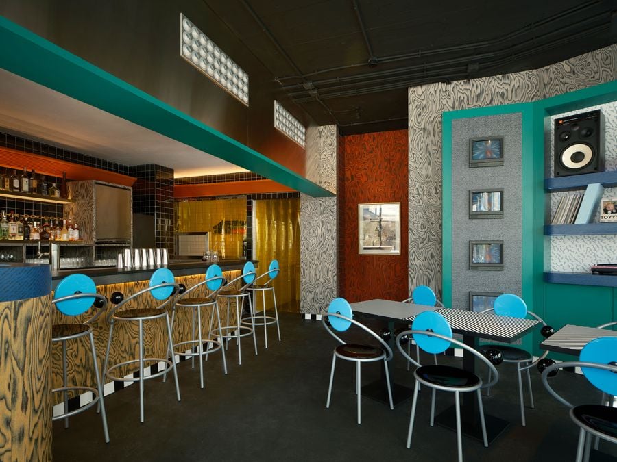 Michele De Lucchi’s First stools line the curved bar, its ochre wood-grain front warmed by concealed lighting, while an amber strip curtain glows towards the kitchen. To the right, striped tables and blue disc-backed chairs, also from Michele De Lucchi’s First series face a rust-red veneered wall. Beside it stands a teal media unit stacked with vintage monitors and speakers.