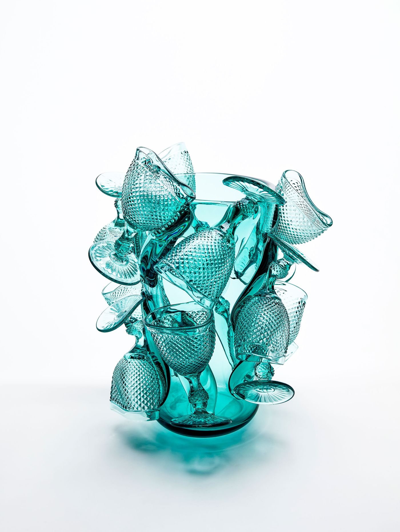 A striking sculptural composition of teal-colored glass goblets, each featuring a textured diamond pattern, emerges from a central, translucent teal vase. This object from Vista Alegre playfully reinterprets traditional glassware, creating a dynamic and artful composition with vibrant color and tactile surfaces.