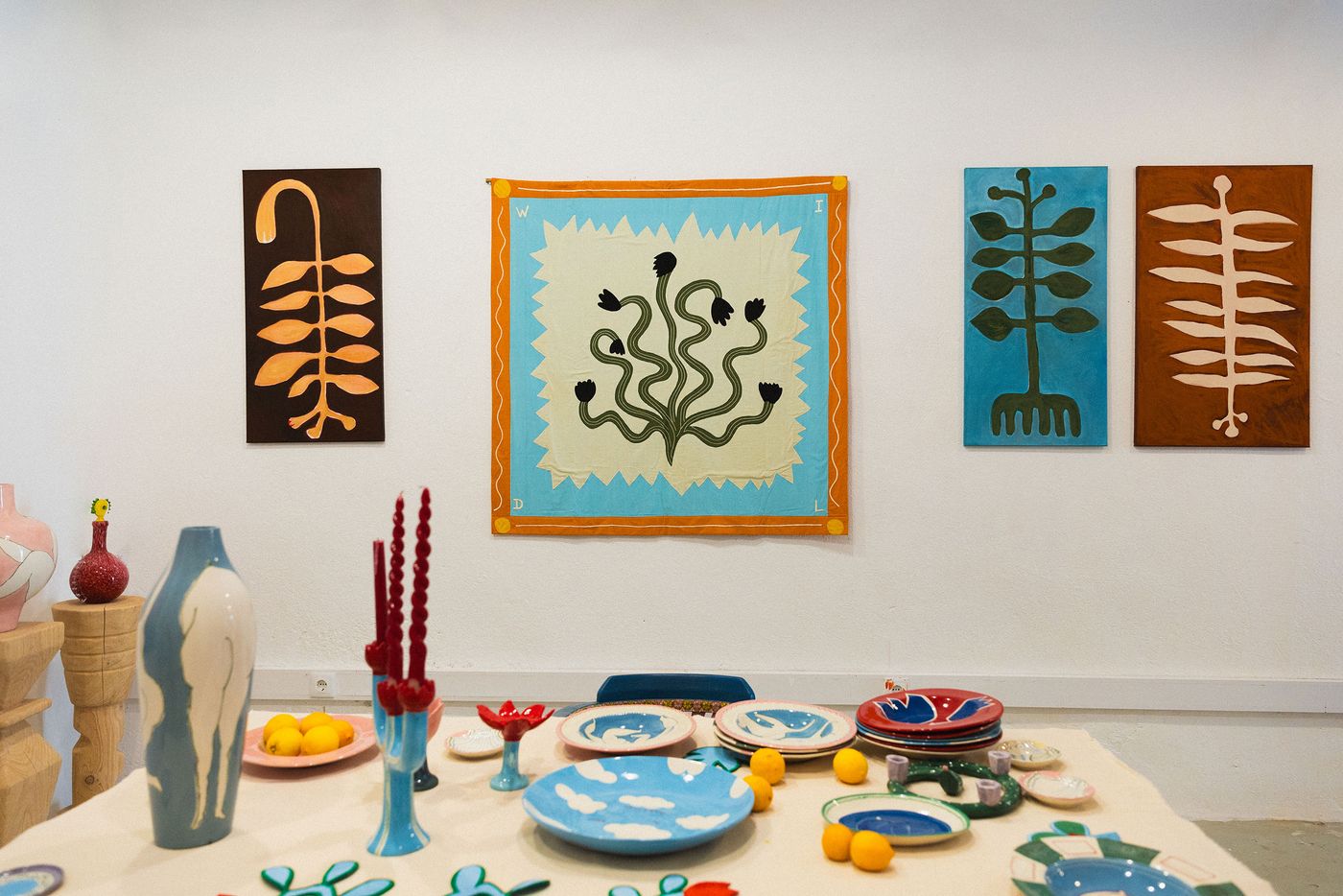A curated exhibition space by Studio Mirante showcasing a collection of vibrant, hand-painted ceramics and wall art. On the table, unique vases and plates in blues, reds, and greens are interspersed with bright lemons. On the white wall, four abstract botanical paintings, varying in color and composition, reflect a cohesive artistic vision with a natural motif. A curated exhibition space by Studio Mirante showcasing a collection of vibrant, hand-painted ceramics and wall art. On the table, unique vases and plates in blues, reds, and greens are interspersed with bright lemons. On the white wall, four abstract botanical paintings, varying in color and composition, reflect a cohesive artistic vision with a natural motif.