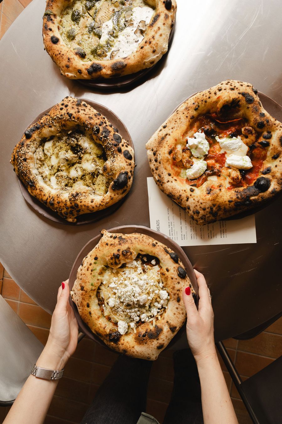 Overhead view of four artisanal pizzas with thick, blistered, charred crusts and assorted cheese, herb and vegetable toppings, arranged on dark ceramic plates across a metallic table at 72H in Athens. A pair of hands with red-painted fingernails holds one plate, adding a candid, convivial touch to the composition.