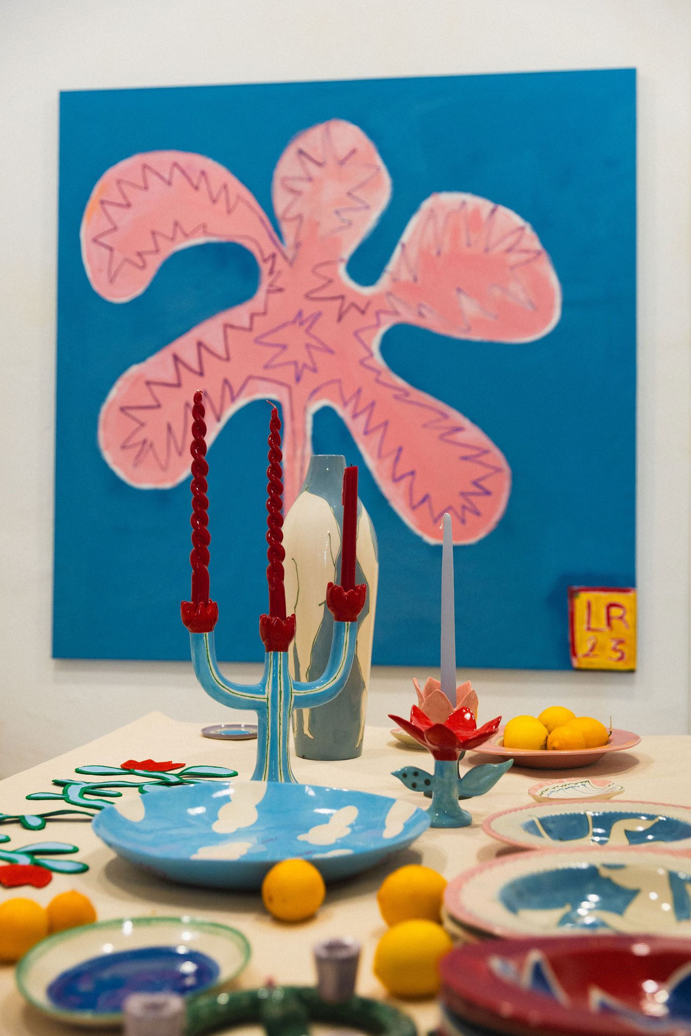 A vibrant tabletop display by Studio Mirante, showcasing eclectic ceramics and playful decor. In the foreground, a blue candelabra with red candles complements a sky-and-cloud patterned plate. A large, abstract pink amoeboid painting with jagged edges dominates the background, set against a bold blue wall, exuding a whimsical, maximalist artistry.