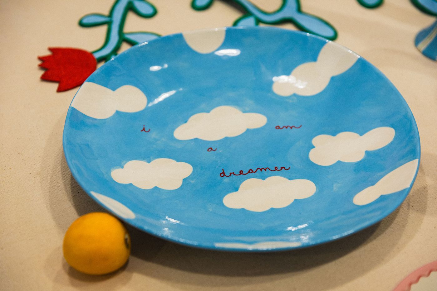 A close-up of a whimsical ceramic plate from Studio Mirante, painted a cheerful sky blue with fluffy white clouds and the handwritten phrase "i am a dreamer." A single bright yellow lemon sits beside it, emphasizing the playful, dreamlike quality and the artisanal charm of the piece.