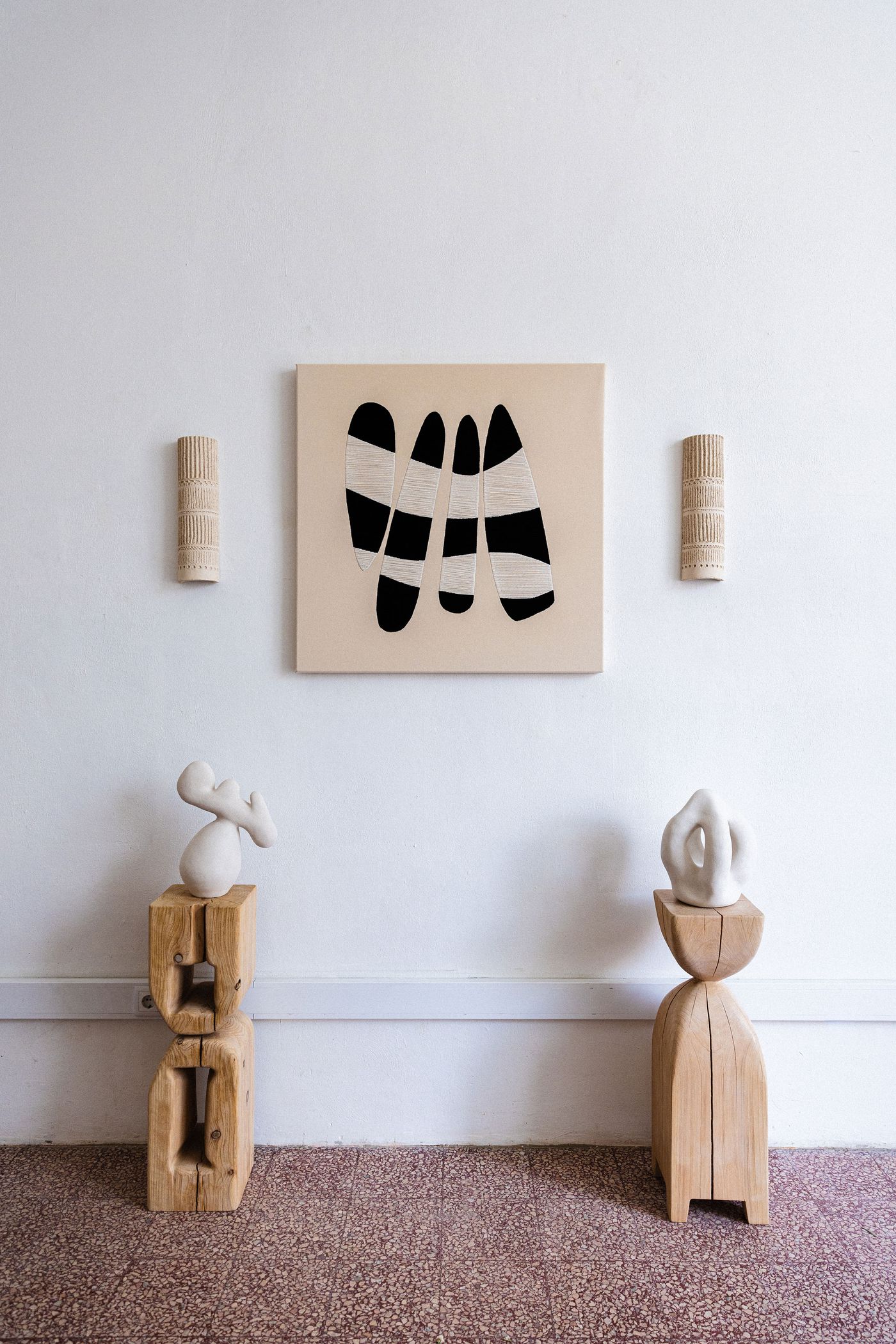 A minimalist gallery setting by Studio Mirante features a central abstract painting with bold black and white organic shapes on a beige canvas. Flanking the painting are two textured, cylindrical wooden wall sconces. Below, two large, abstract white ceramic sculptures rest on rustic wooden plinths, creating a harmonious interplay of form, texture, and natural materials. A minimalist gallery setting by Studio Mirante features a central abstract painting with bold black and white organic shapes on a beige canvas. Flanking the painting are two textured, cylindrical wooden wall sconces. Below, two large, abstract white ceramic sculptures rest on rustic wooden plinths, creating a harmonious interplay of form, texture, and natural materials.
