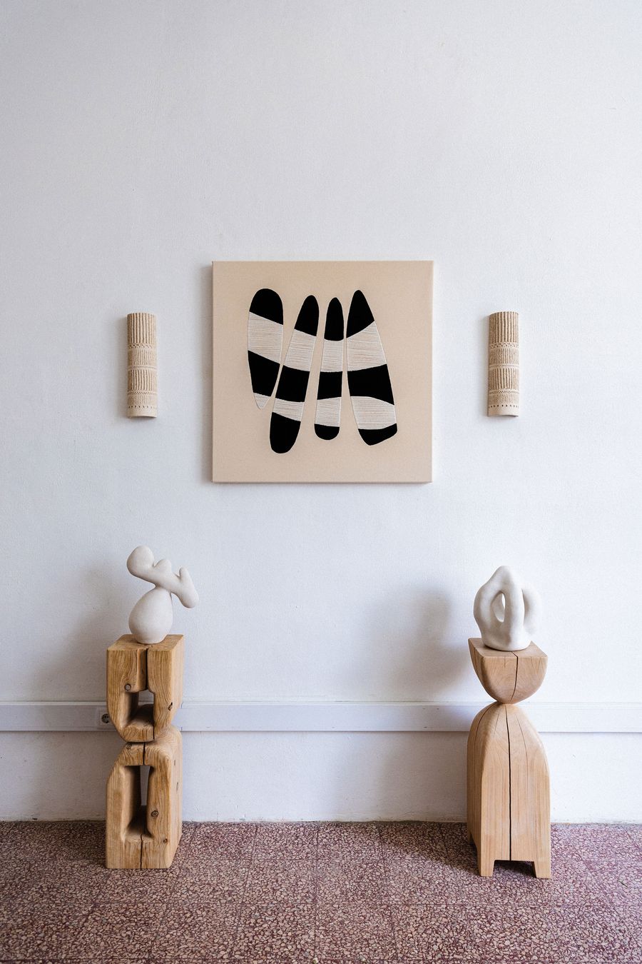 A minimalist gallery setting by Studio Mirante features a central abstract painting with bold black and white organic shapes on a beige canvas. Flanking the painting are two textured, cylindrical wooden wall sconces. Below, two large, abstract white ceramic sculptures rest on rustic wooden plinths, creating a harmonious interplay of form, texture, and natural materials.