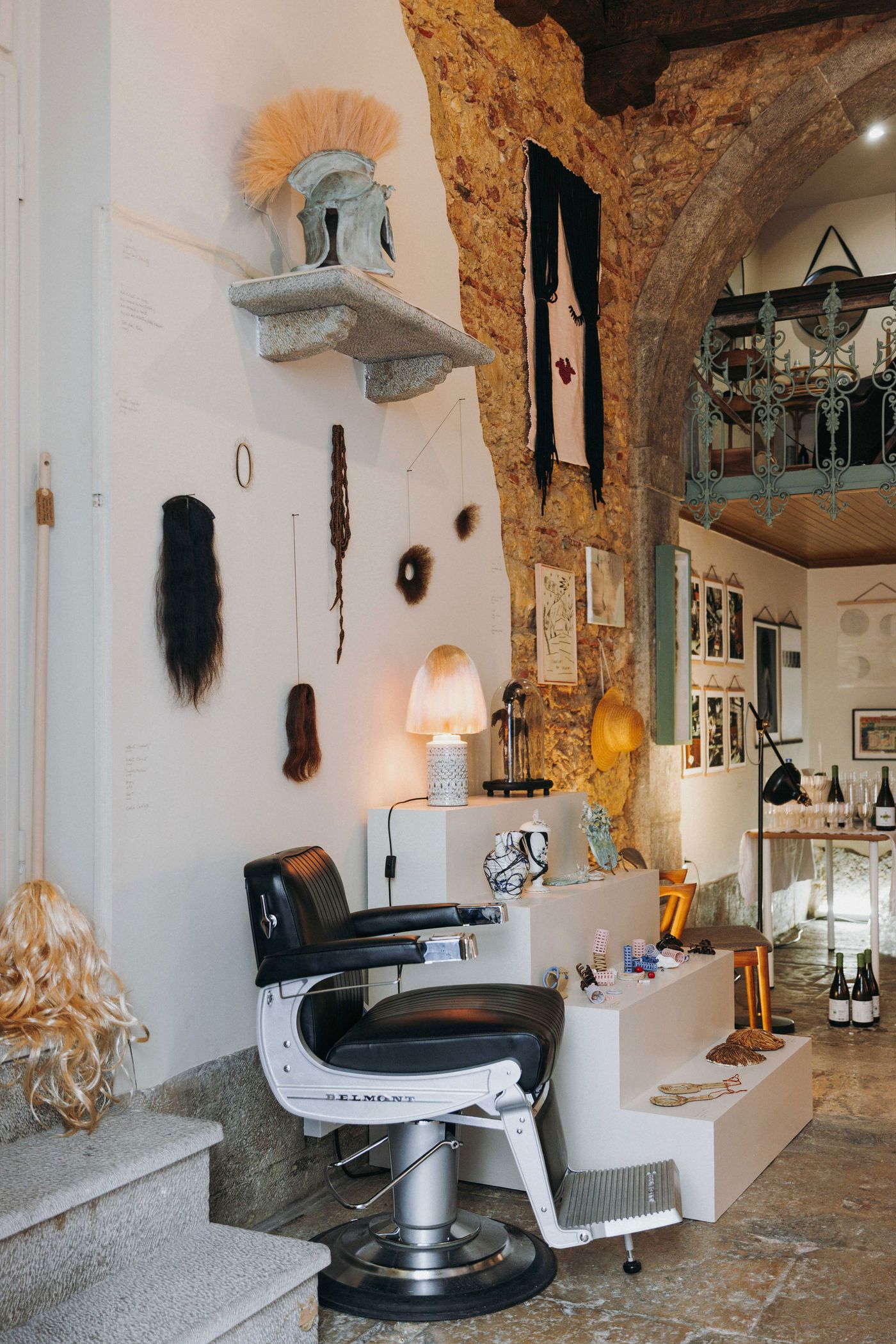 The “Tangled Narratives” exhibition at Thiago Novaes' hair studio which merges industrial and eclectic aesthetics within a stone-walled space. A vintage black leather barber chair sits before a white display wall adorned with various hair extensions and artistic pieces. Stone shelves and an exposed archway frame the scene, creating a unique, almost curio-shop ambiance.