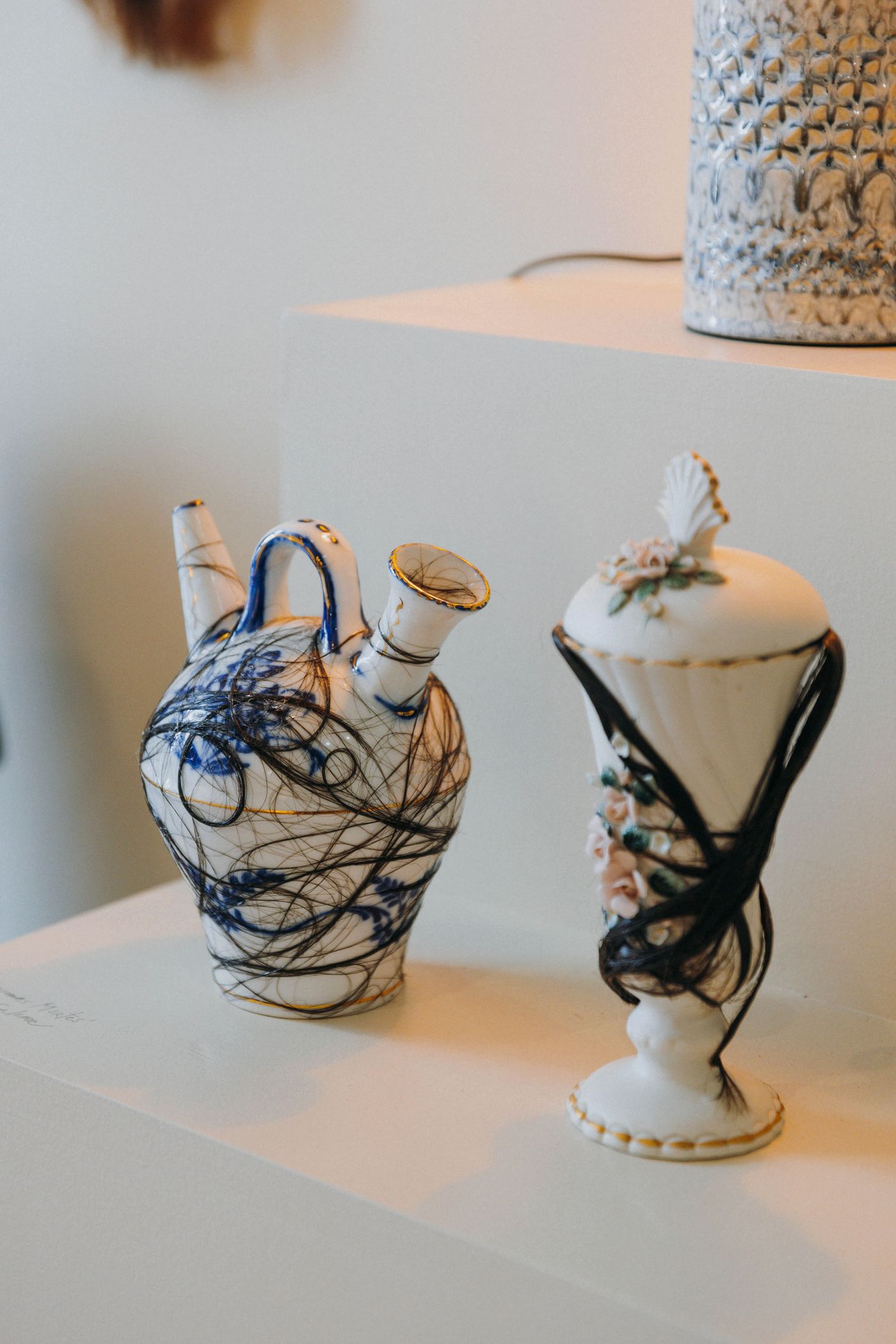 Two unusual ceramic pieces from the “Tangled Narratives” exhibition are displayed in Thiago Novaes' hair salon. On the left, a white pitcher with blue abstract markings is adorned with dark, tangled hair strands. On the right, a lidded urn with delicate floral embellishments also features dark hair, highlighting a surreal and provocative exploration of hair as an artistic medium.