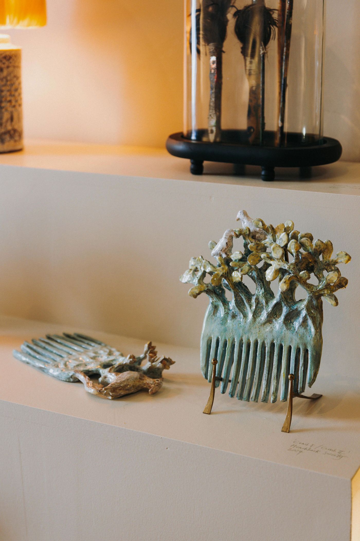 Two sculptural hair combs from the "Tangled Narratives" exhibition at Thiago Novaes' hair salon are displayed on a clean, light surface. Each comb features intricate, organic adornments resembling moss-covered branches, showcasing a unique fusion of functional objects with natural, artistic elements, embodying a biophilic design philosophy.