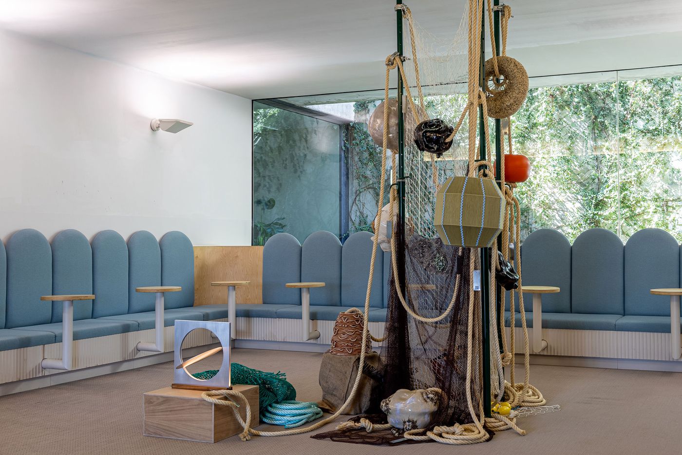 A wide view of LACS Santos' "The Tide Line Exhibition" captures an immersive, nature-inspired installation. Thick ropes, fishing nets, and marine elements like buoys cascade from central poles, grounded by a collection of objects including a bird-shaped sculpture. The textural, rustic art piece contrasts with the clean-lined, modern blue seating area and large windows revealing vibrant greenery. A wide view of LACS Santos' "The Tide Line Exhibition" captures an immersive, nature-inspired installation. Thick ropes, fishing nets, and marine elements like buoys cascade from central poles, grounded by a collection of objects including a bird-shaped sculpture. The textural, rustic art piece contrasts with the clean-lined, modern blue seating area and large windows revealing vibrant greenery.