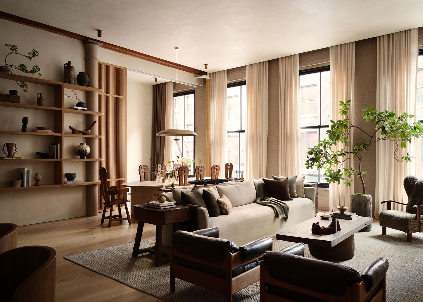 A stylish, airy living room featuring large windows, a comfortable sofa, wooden shelves with decor, and a dining area with elegant chairs. A stylish, airy living room featuring large windows, a comfortable sofa, wooden shelves with decor, and a dining area with elegant chairs.
