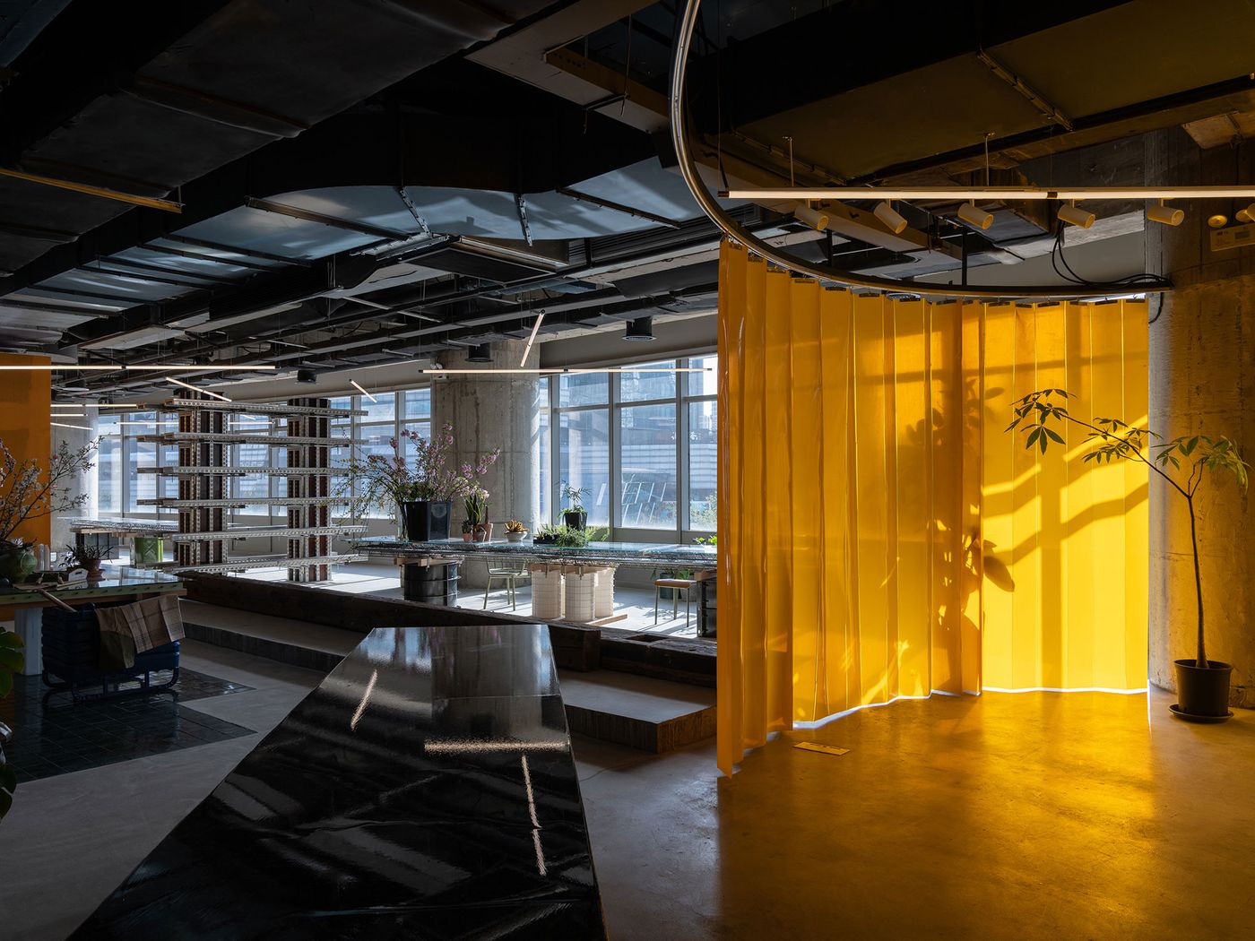 A sun-drenched area of 889 GLO Art Space is defined by a vivid yellow curtain, creating a dramatic interplay of light and shadow. The industrial aesthetic is evident in exposed concrete columns and ceilings. A sleek, dark marble counter in the foreground adds a touch of refinement to the otherwise raw and adaptable exhibition space.