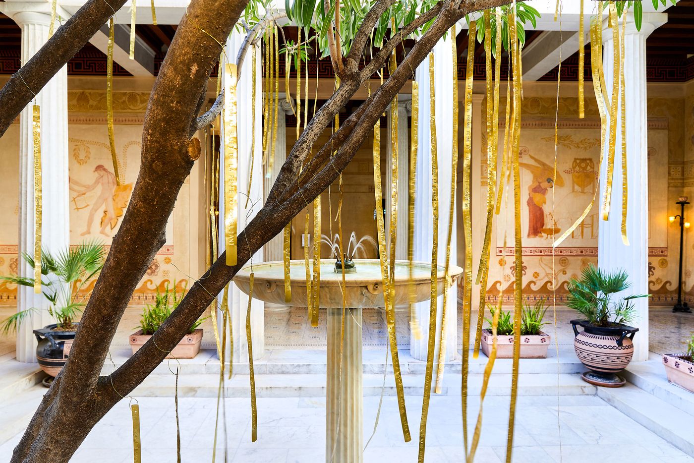 A peaceful courtyard at Villa Kerylos with a central fountain. A tree with thin leaves and a mossy trunk stands next to the fountain, its branches adorned with long, golden strips of metal. The neo-vernacular walls feature faded murals depicting figures and motifs from classical mythology, creating a biophilic and artful space. A peaceful courtyard at Villa Kerylos with a central fountain. A tree with thin leaves and a mossy trunk stands next to the fountain, its branches adorned with long, golden strips of metal. The neo-vernacular walls feature faded murals depicting figures and motifs from classical mythology, creating a biophilic and artful space.