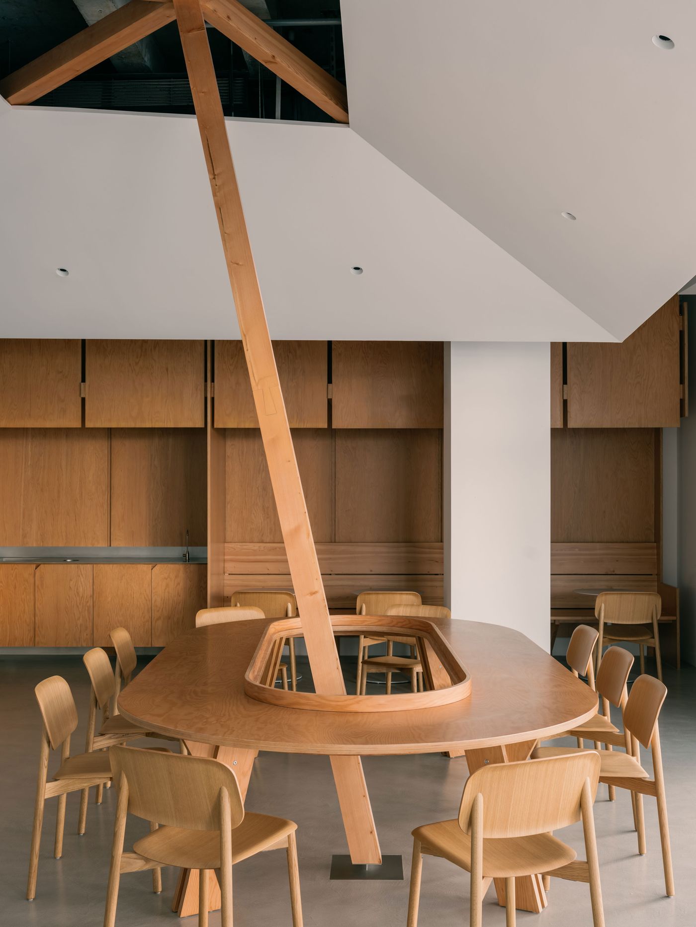 A close-up of the central communal table shows its unique construction—an oval form punctured by a structural wooden beam rising to meet the angular ceiling with sculptural precision.
