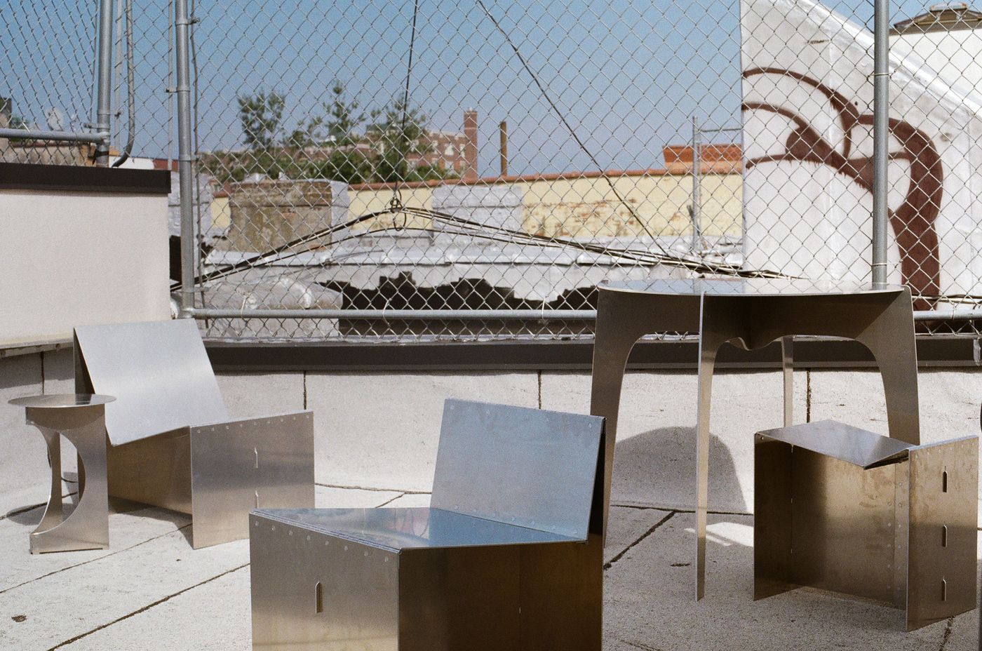 A collection of industrial-style metal furniture is set on a rooftop terrace, contrasting with the gritty urban background. The pieces—two chairs, a stool, and a table—are made from folded and riveted sheet metal, their reflective surfaces catching the light. The composition highlights the geometric forms and functional nature of the pieces, juxtaposing their polished finish against the chain-link fence and weathered buildings, creating a raw, brutalist aesthetic.
