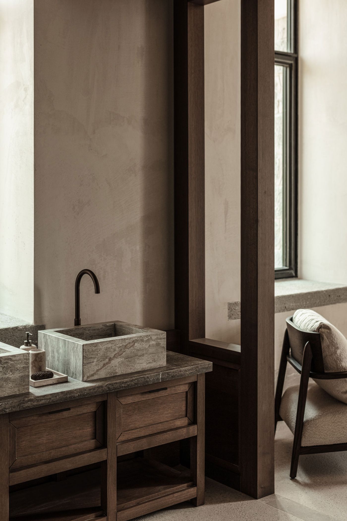 A serene minimalist bathroom features a textured stone vessel sink and a matte black faucet, set against a backdrop of muted, earthy plaster walls. Dark timber accents, including a framed partition and window, create a striking contrast, enhancing the space's tranquil and sophisticated atmosphere, characteristic of a contemporary Japandi aesthetic. A serene minimalist bathroom features a textured stone vessel sink and a matte black faucet, set against a backdrop of muted, earthy plaster walls. Dark timber accents, including a framed partition and window, create a striking contrast, enhancing the space's tranquil and sophisticated atmosphere, characteristic of a contemporary Japandi aesthetic.