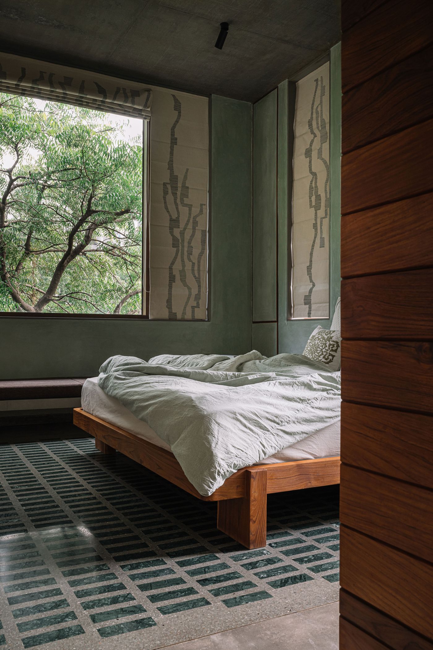 A serene bedroom features a low-profile wooden bed with light grey bedding. The walls are a calming sage green, accented by two tall fabric panels with abstract black and white patterns. A large window offers a view of dense tree foliage, while a dark green and grey patterned tiled floor adds subtle visual interest, creating a tranquil and earthy retreat.