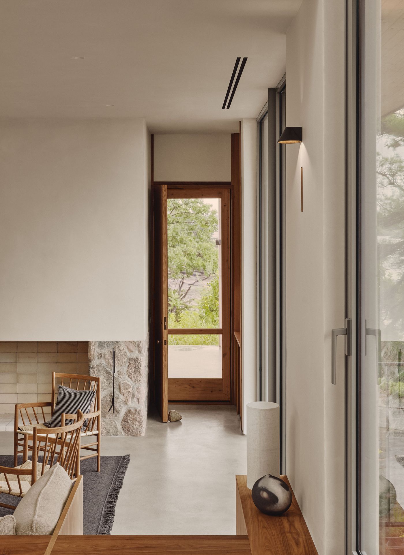 Bright, modern interior with a stone fireplace, wooden chairs, and a view of greenery through large windows and an open door. Bright, modern interior with a stone fireplace, wooden chairs, and a view of greenery through large windows and an open door.
