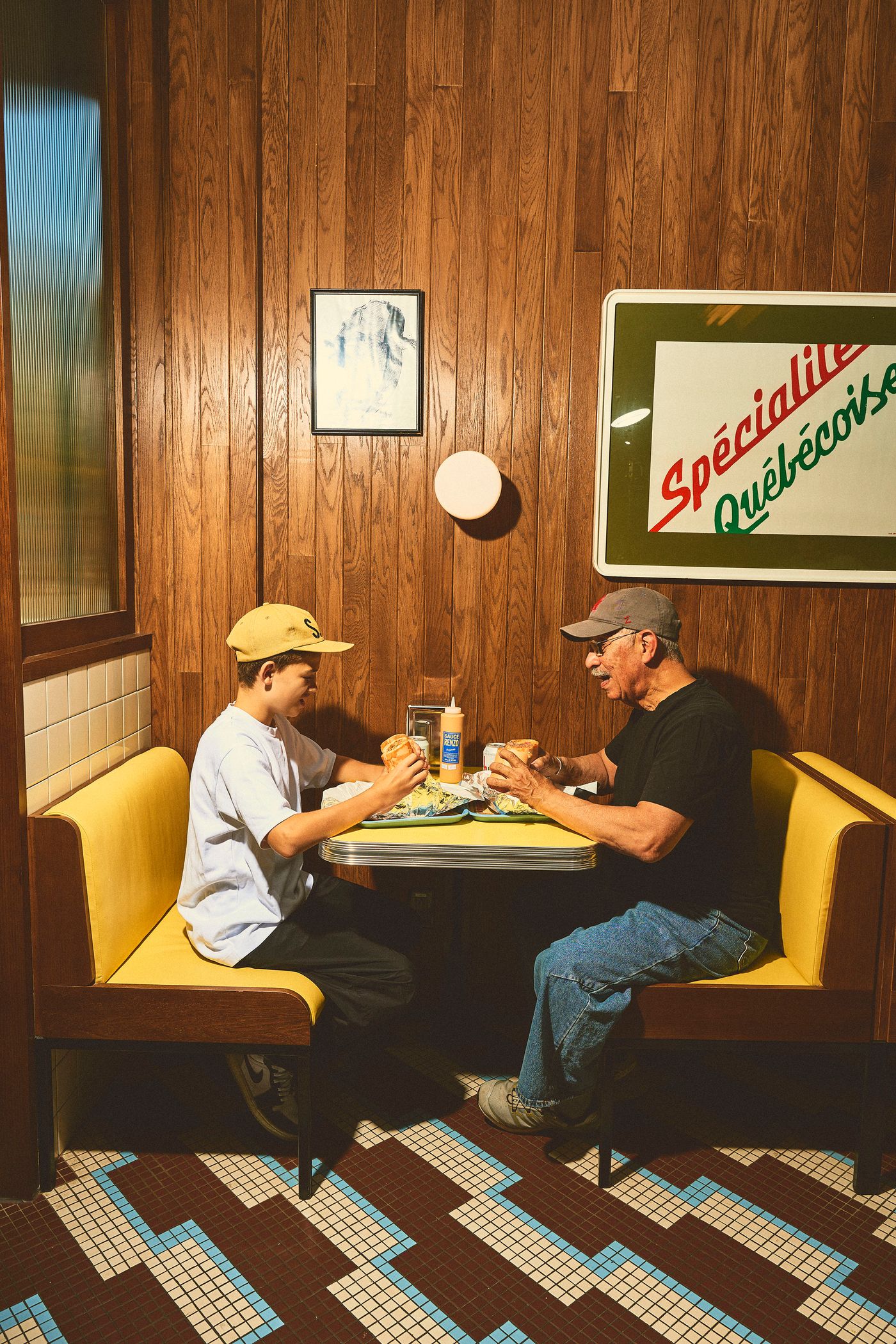 An older man and a young boy sit opposite each other in a booth at Renzo Sandwich. Yellow banquettes and wood-paneled walls create a retro atmosphere. The table holds sandwiches and condiments. Above, a sign reads "Spécialité Québécoise," hinting at local flavors. The tiled floor adds a classic diner touch.