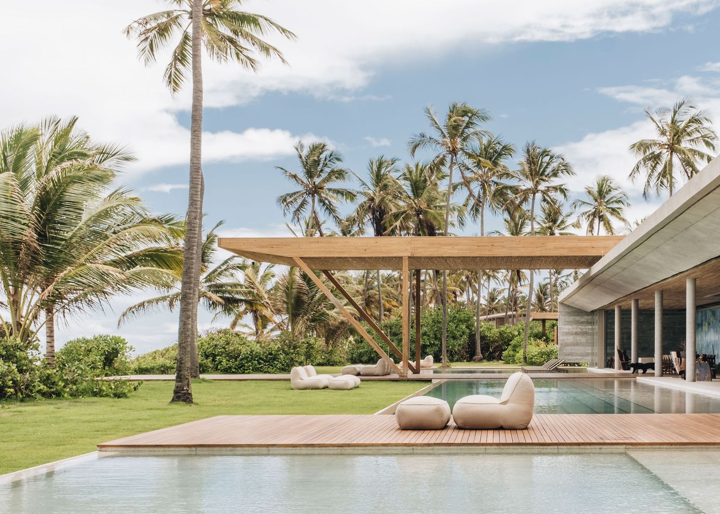 Modern villa with a wooden patio overlooking a pool, surrounded by palm trees and lush greenery under a bright blue sky.