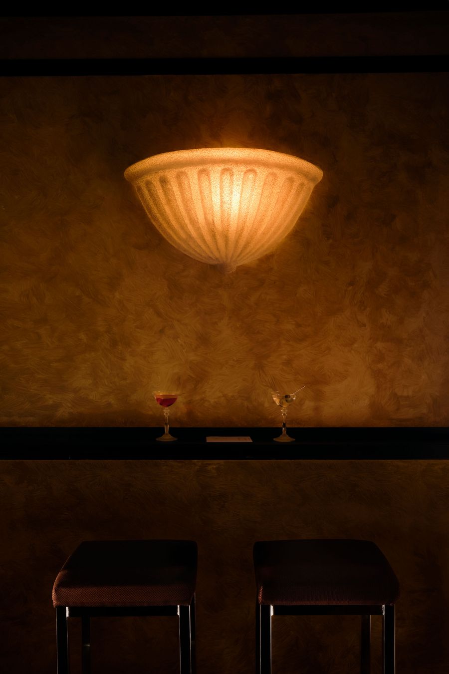 An intimate vignette at The Roses of Elagabalus captures a classic, fluted wall sconce emitting a warm, diffused light onto a textured, amber-gold wall. Below, two elegant cocktail glasses, one with a red drink and the other clear, rest on a dark shelf above two barstools, creating a sophisticated and inviting ambiance. An intimate vignette at The Roses of Elagabalus captures a classic, fluted wall sconce emitting a warm, diffused light onto a textured, amber-gold wall. Below, two elegant cocktail glasses, one with a red drink and the other clear, rest on a dark shelf above two barstools, creating a sophisticated and inviting ambiance.