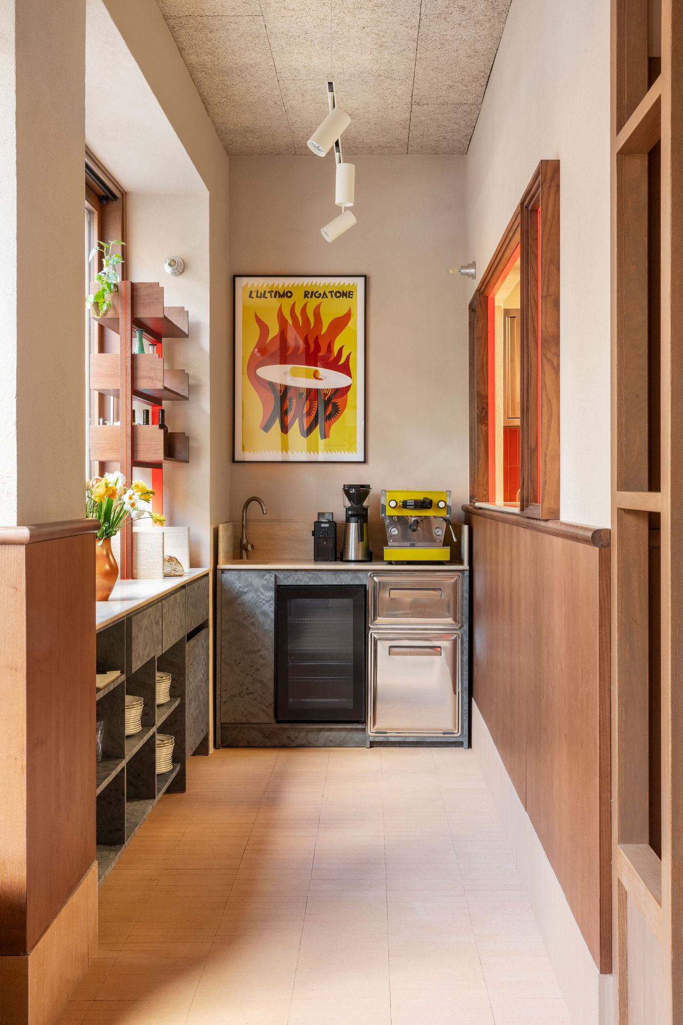 A dynamic service area within Santo Palato by Naessi Studio, combining functionality with playful accents. A vibrant, graphic poster hangs above a compact counter with a bright yellow espresso machine. Wooden wainscoting and shelves line the walls, while exposed track lighting illuminates the practical yet characterful space, reflecting a modern Italian sensibility. A dynamic service area within Santo Palato by Naessi Studio, combining functionality with playful accents. A vibrant, graphic poster hangs above a compact counter with a bright yellow espresso machine. Wooden wainscoting and shelves line the walls, while exposed track lighting illuminates the practical yet characterful space, reflecting a modern Italian sensibility.