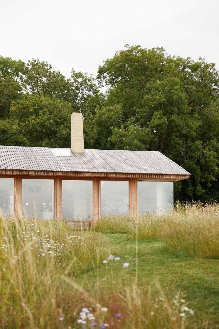 The Maker's Barn, a contemporary structure by Hutch Design, is enveloped by a vibrant, wild meadow transitioning to manicured lawn. Its low-slung form features a horizontal timber-clad roof and a wall of large, clear windows framed in light wood, reflecting the sky and trees, while a distinctive pale chimney anchors the building, showcasing a seamless blend of modern and natural elements. The Maker's Barn, a contemporary structure by Hutch Design, is enveloped by a vibrant, wild meadow transitioning to manicured lawn. Its low-slung form features a horizontal timber-clad roof and a wall of large, clear windows framed in light wood, reflecting the sky and trees, while a distinctive pale chimney anchors the building, showcasing a seamless blend of modern and natural elements.