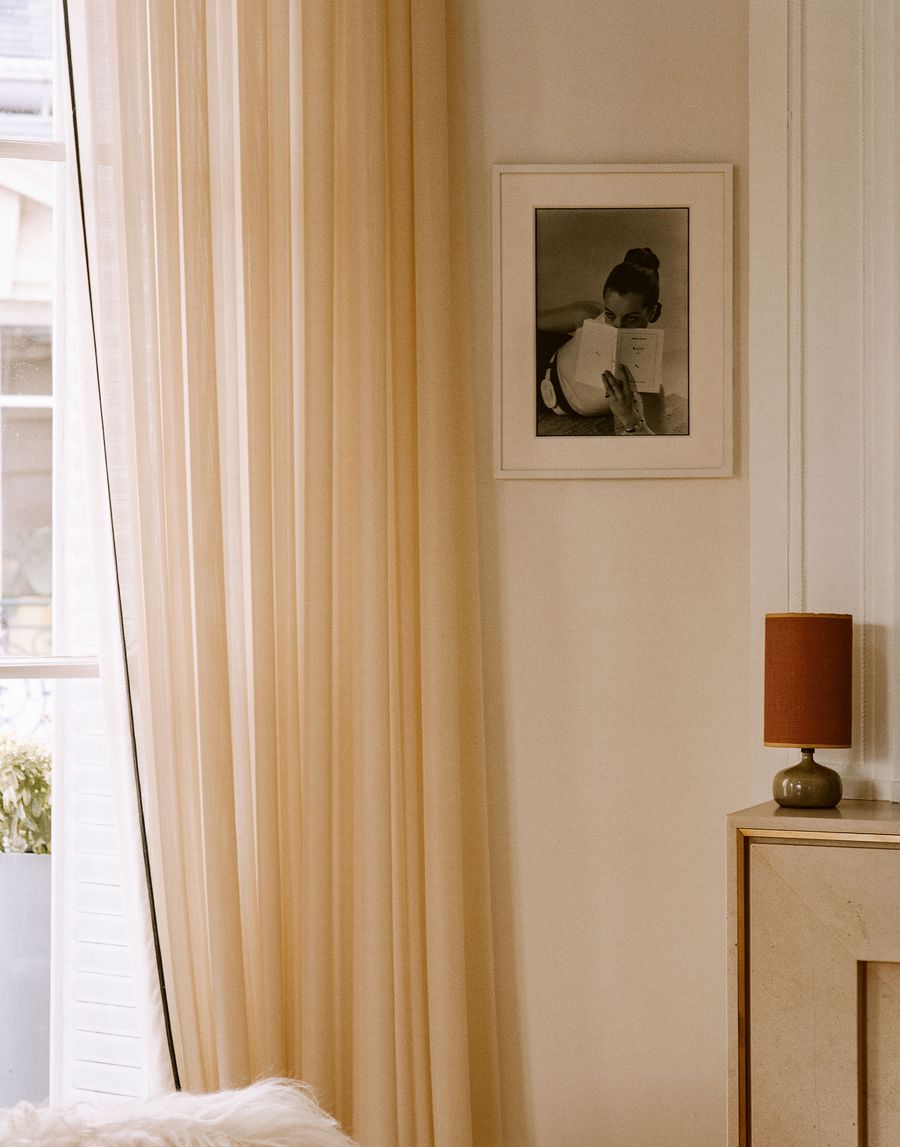 A warm, serene corner features a cream-colored sheer curtain softly diffusing natural light from a large window. A framed black and white photograph hangs on the wall, subtly complementing the minimalist aesthetic and the rustic-toned table lamp on a light marble console. A warm, serene corner features a cream-colored sheer curtain softly diffusing natural light from a large window. A framed black and white photograph hangs on the wall, subtly complementing the minimalist aesthetic and the rustic-toned table lamp on a light marble console.