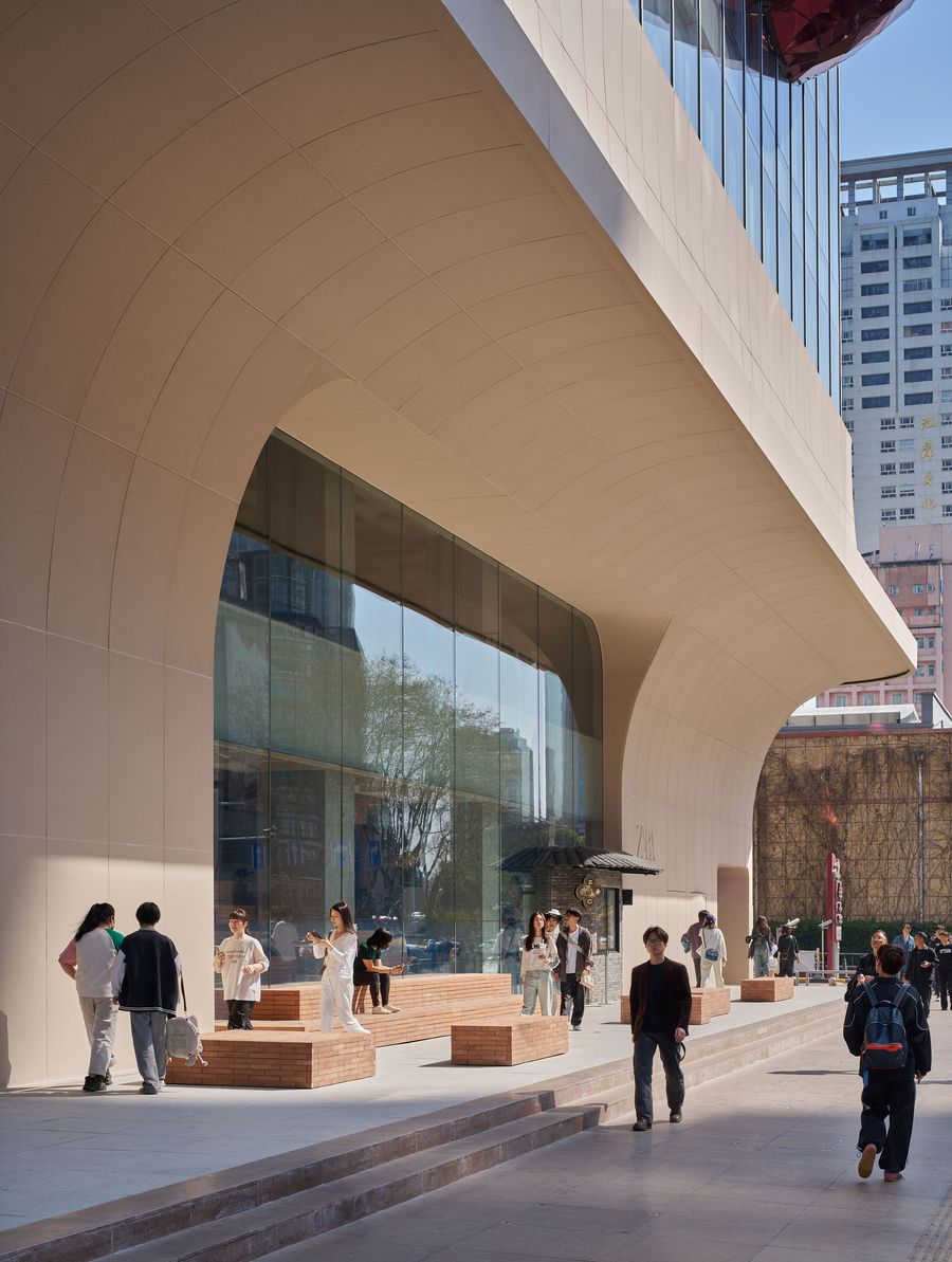 A modern building with a curved facade features groups of people seated on benches and walking along a wide, paved area. A modern building with a curved facade features groups of people seated on benches and walking along a wide, paved area.
