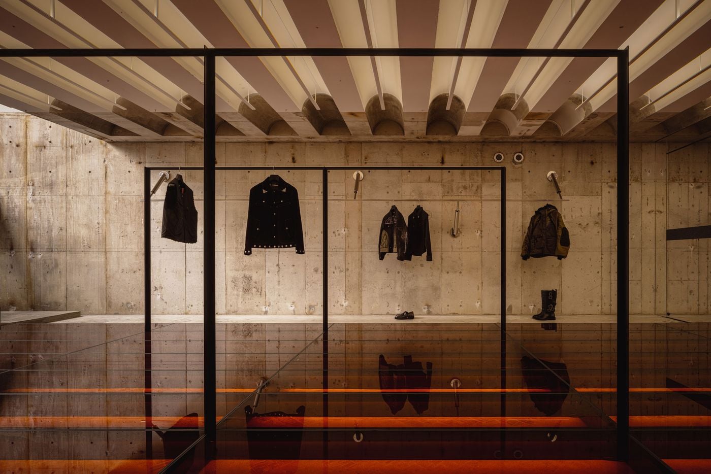 Black metal rails form a series of large rectangular frames across the basement, setting sparse garments against unfinished concrete walls. Below, the reflective glass floor captures warm red illumination from beneath, while the linear ceiling treatment above creates a striking dialogue between repetition, transparency and the building’s rough Brutalist fabric.