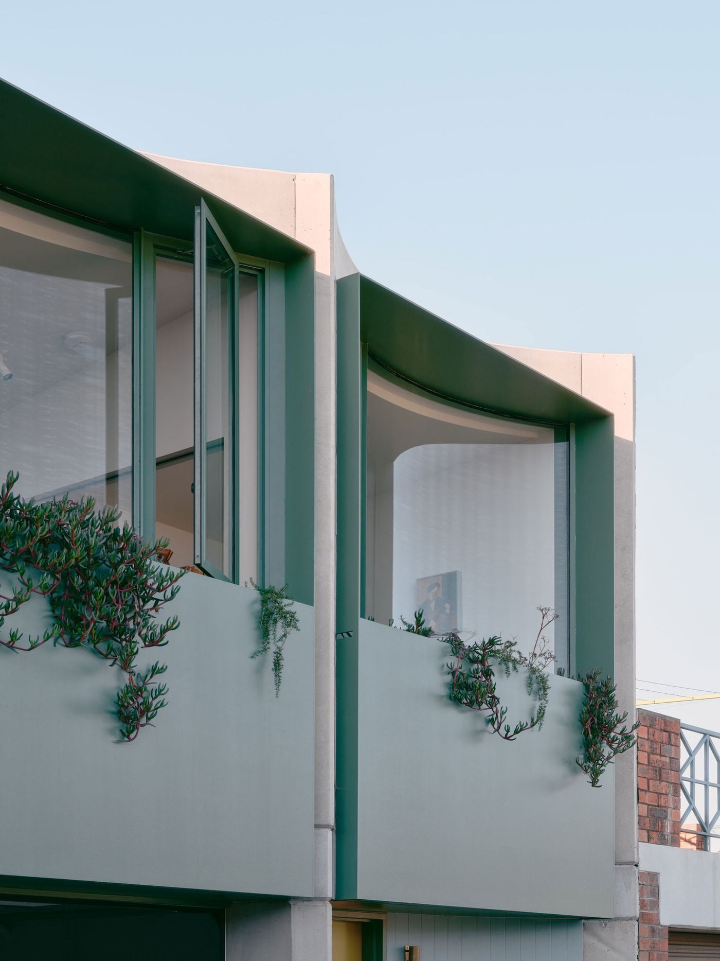 A low-angle detail emphasises the sinuous geometry of Denj Lane’s upper façade, where curved glazing and deep green metal reveals sit between pale concrete fins. Openable windows and trailing succulents animate the surface, while the alternating concave bays give the compact addition an unexpectedly expressive profile. A low-angle detail emphasises the sinuous geometry of Denj Lane’s upper façade, where curved glazing and deep green metal reveals sit between pale concrete fins. Openable windows and trailing succulents animate the surface, while the alternating concave bays give the compact addition an unexpectedly expressive profile.