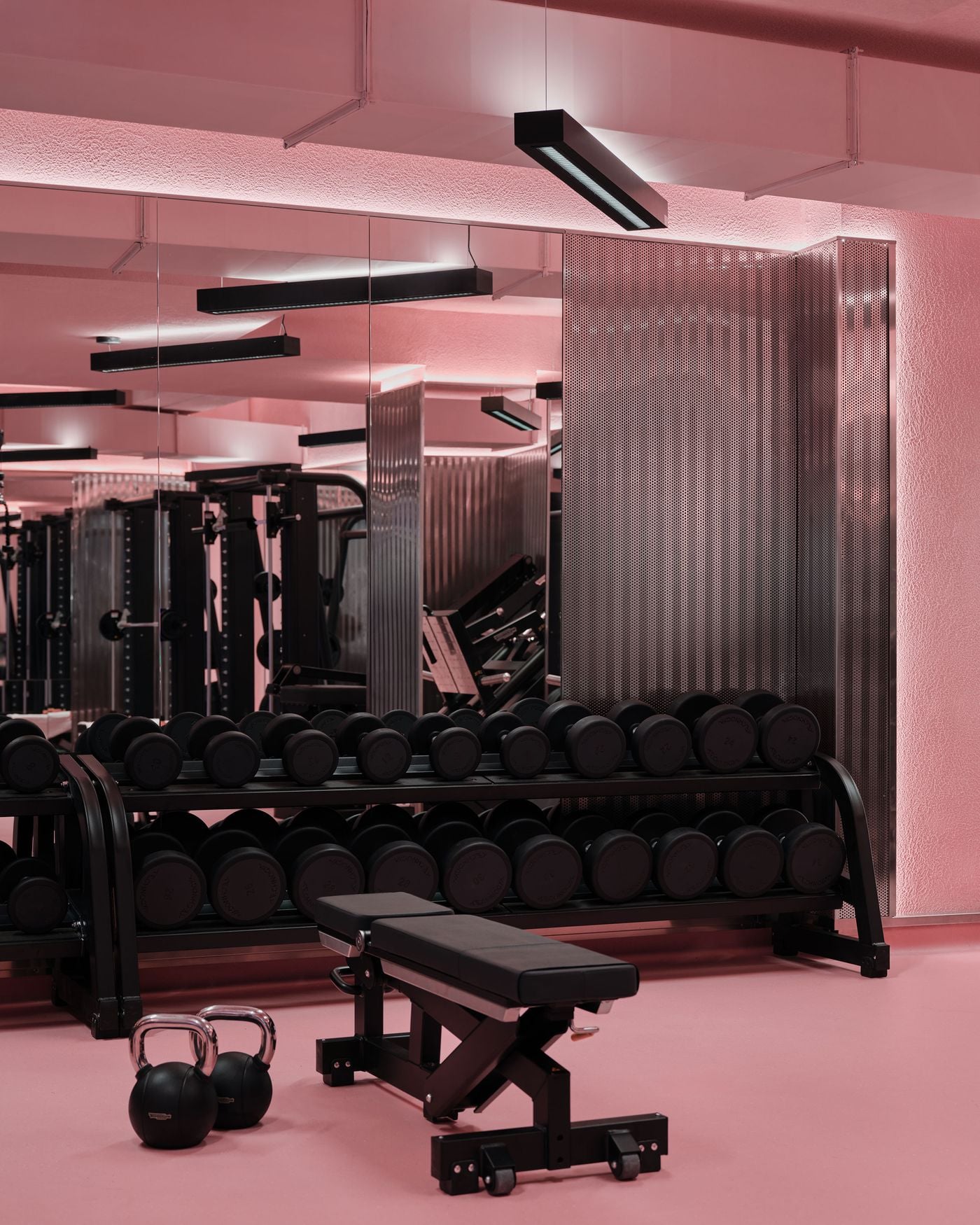 The open training floor is washed in cotton-candy pink, offset by black gym equipment, mirrored walls and perforated metallic partitions. Suspended linear lights and reflective surfaces heighten the graphic effect, while the restrained equipment palette allows the colour treatment to define the room’s bold, retro-futurist identity. The open training floor is washed in cotton-candy pink, offset by black gym equipment, mirrored walls and perforated metallic partitions. Suspended linear lights and reflective surfaces heighten the graphic effect, while the restrained equipment palette allows the colour treatment to define the room’s bold, retro-futurist identity.