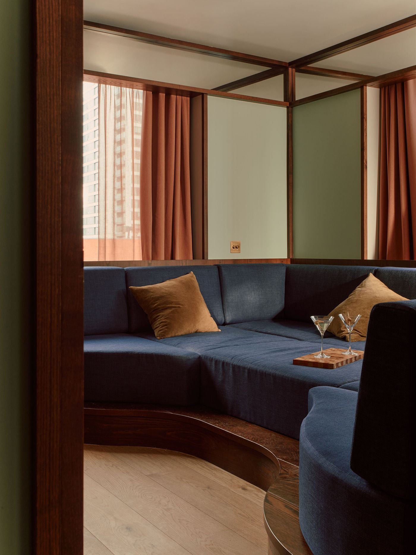 The oversized blue sofa pit turns the guest room into a distinctly social setting, recalling the sunken conversation areas and modular “playpens” of 1960s interiors. Dark walnut edging traces its curving form, while sage-green walls, rust-coloured curtains and soft daylight keep Holloway Li’s nostalgic reference feeling contemporary. The oversized blue sofa pit turns the guest room into a distinctly social setting, recalling the sunken conversation areas and modular “playpens” of 1960s interiors. Dark walnut edging traces its curving form, while sage-green walls, rust-coloured curtains and soft daylight keep Holloway Li’s nostalgic reference feeling contemporary.
